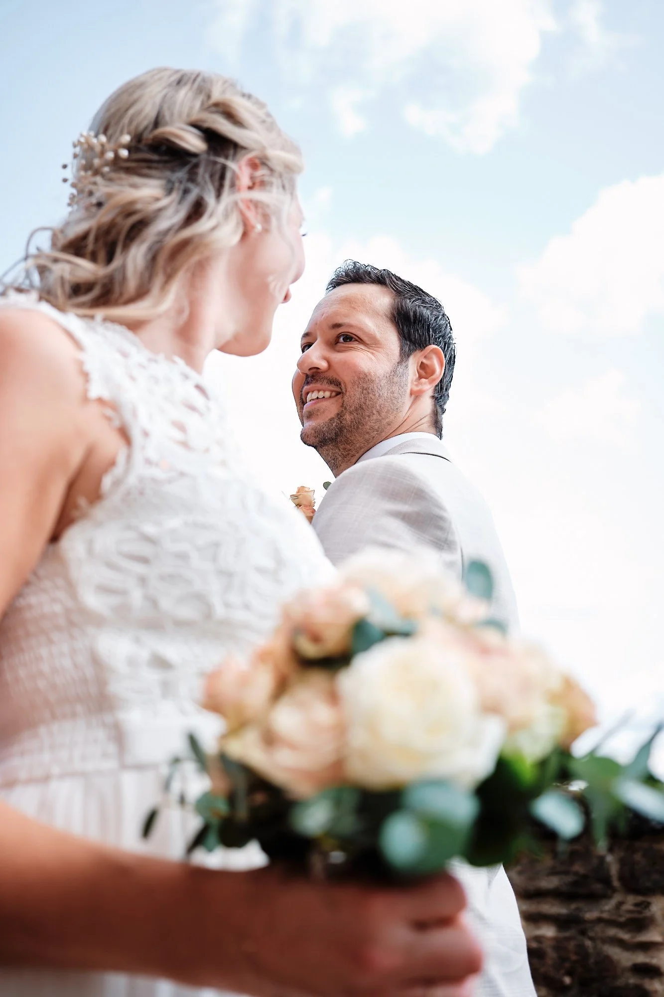 Portrait eines Bräutigams beim Paarshooting: Bräutigam blickt lächelnd zur Braut, Blumenbouquet im Vordergrund, blauer Himmel
