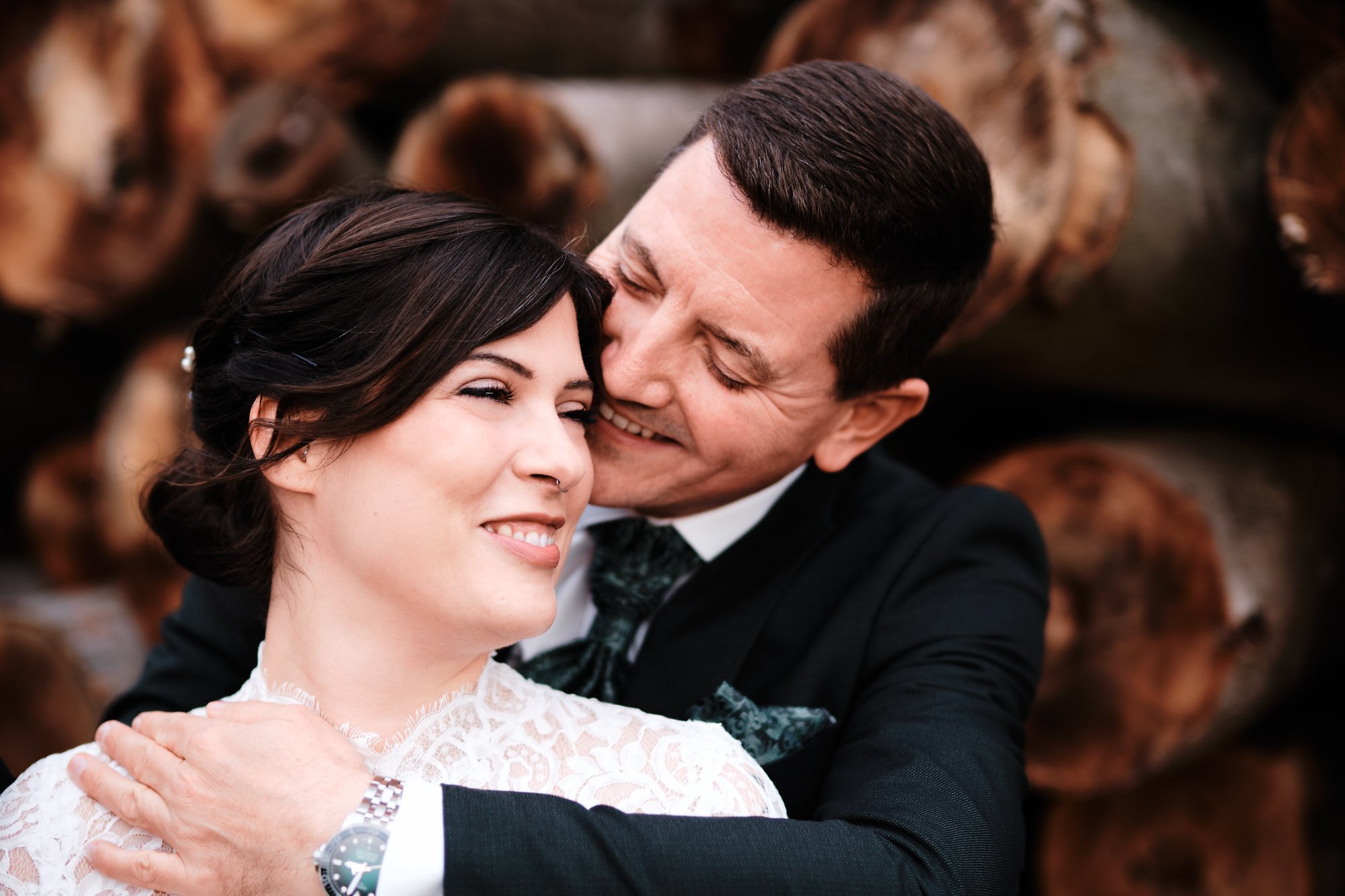 Romantische Hochzeitsaufnahme: Bräutigam umarmt Braut von hinten, lächeln, elegante Hochzeitskleidung, innige Nähe vor Holz