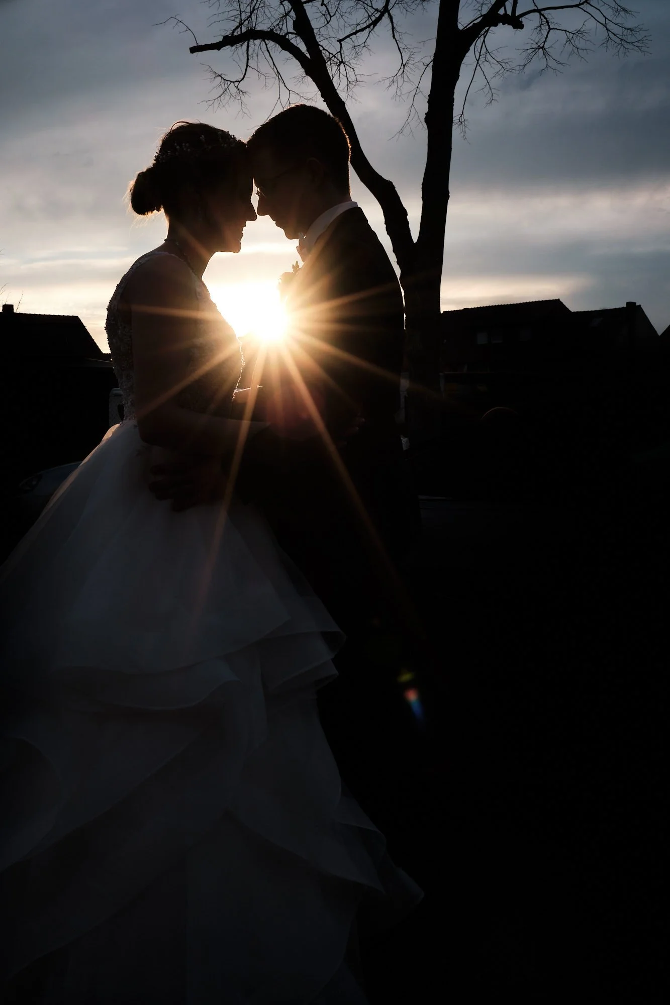 Silhouette eines Hochzeitspaares in der Abendsonne, Braut und Bräutigam berühren sich, Sonnenstrahlen und Baum im Hintergrund.