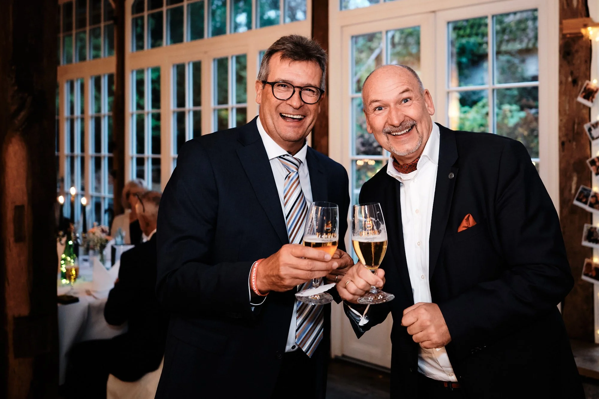 Ausgelassene Feierstimmung auf einer Hochzeit: Zwei Männer in Anzügen stoßen mit Champagnergläsern an und lachen herzlich.