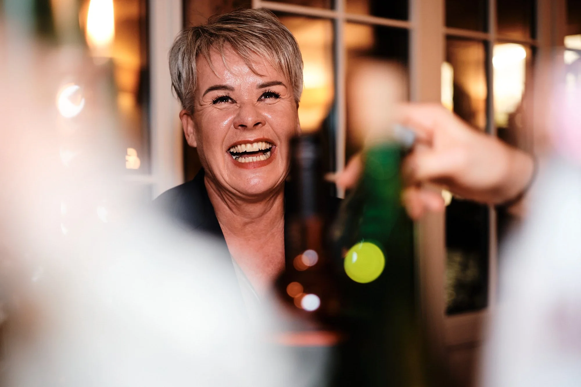 Stimmungsvoller Moment. Portrait einer lachenden Frau am Tisch auf einer Hochzeit: lachend, nah an Getränkeflaschen, gesellige Feier