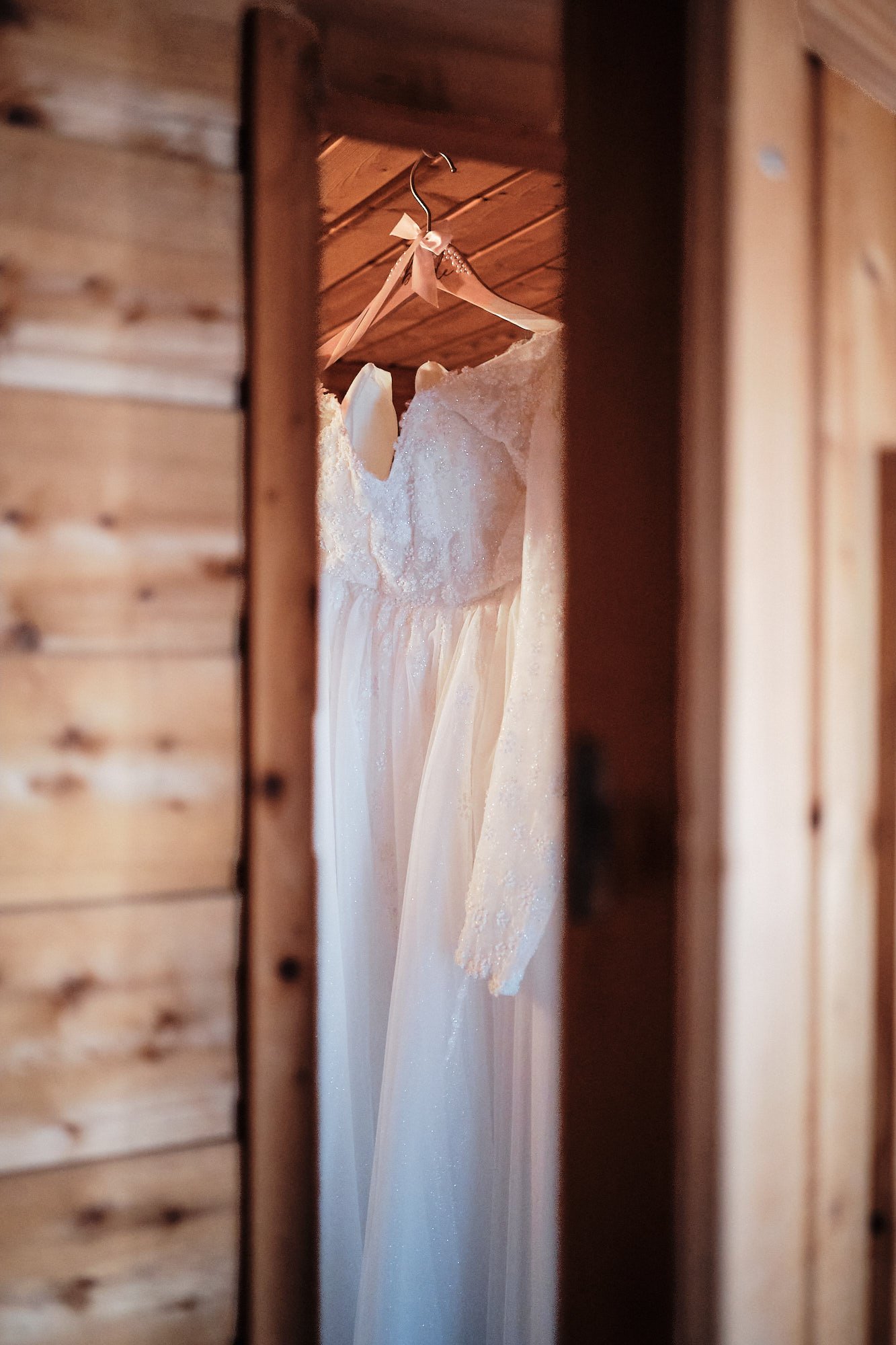 Detailbild eines Hochzeitskleids: Weißes Brautkleid hängt an einem Bügel, durch Türspalt fotografiert.