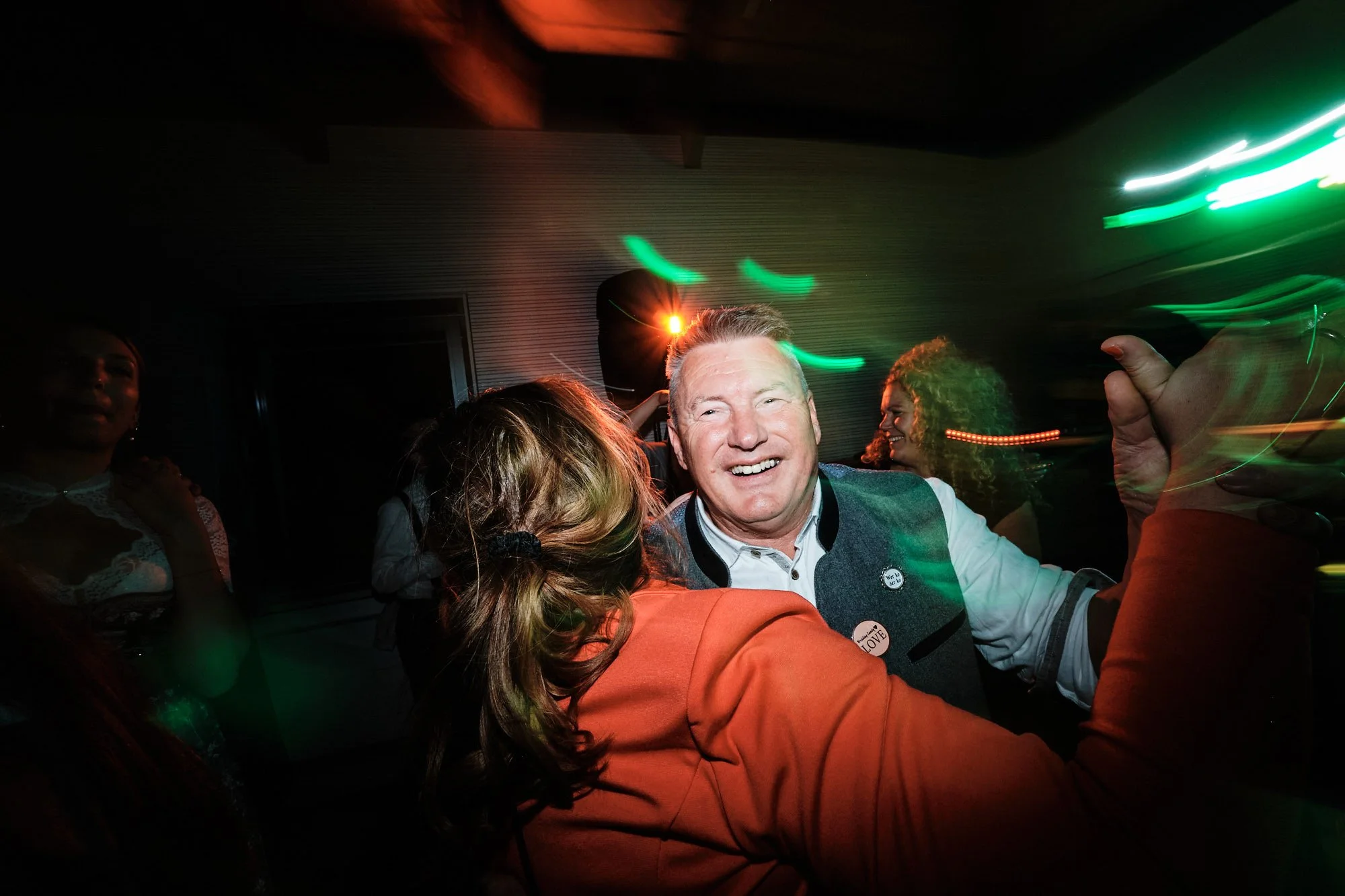 Hochzeitsparty: Bräutigam lächelnd zwischen tanzenden Gästen unter farbigen Lichteffekten.