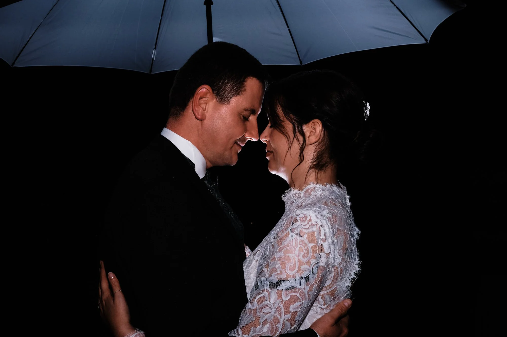 Ruhiges Portrait eines Paares unter Regenschirm in der Nacht – Braut in Spitzenkleid, Bräutigam im Anzug, innige Nähe.