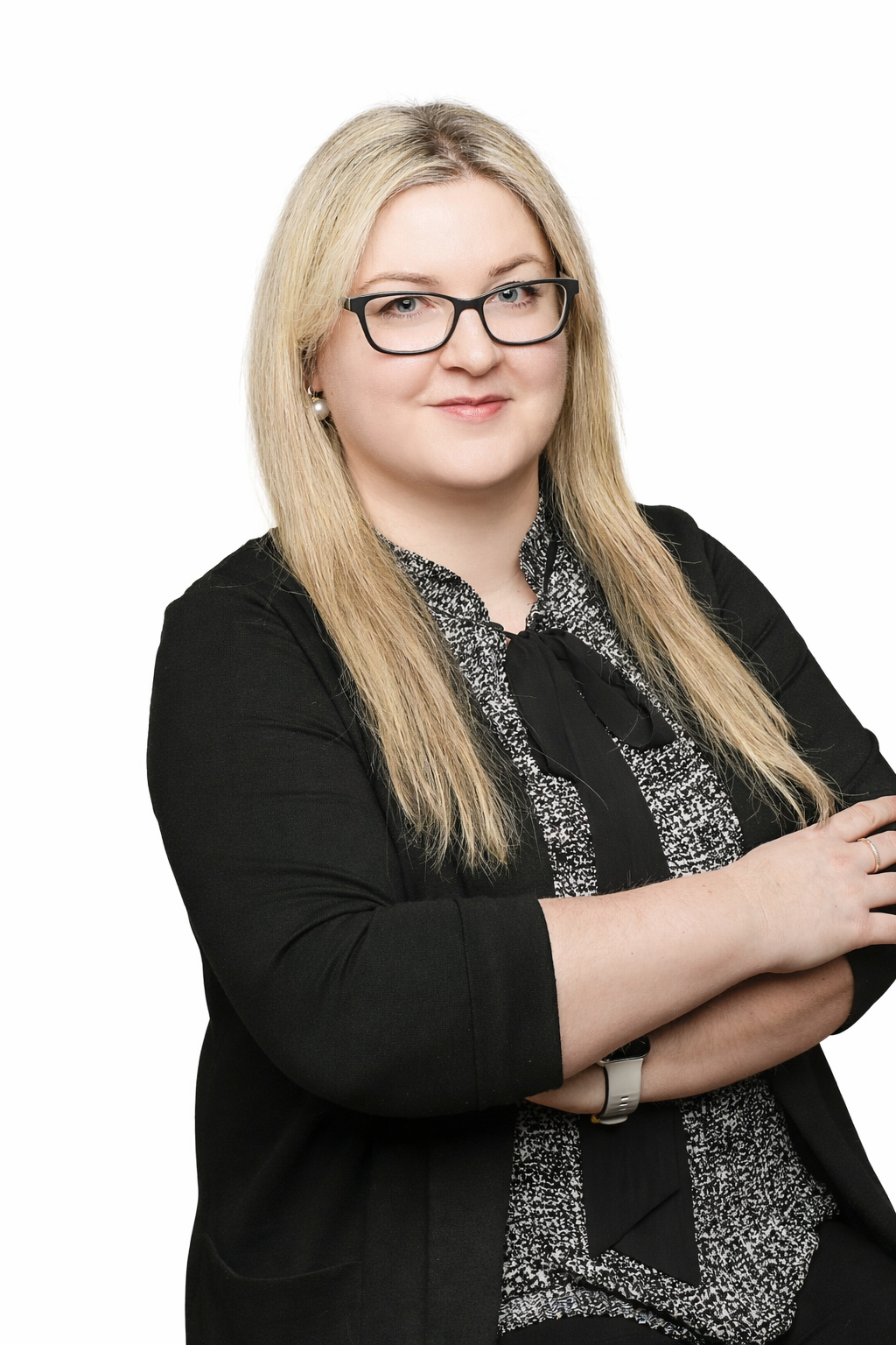 A woman with shoulder-length blonde hair, wearing a white shirt and black blazer, standing in front of a plain light-colored wall.