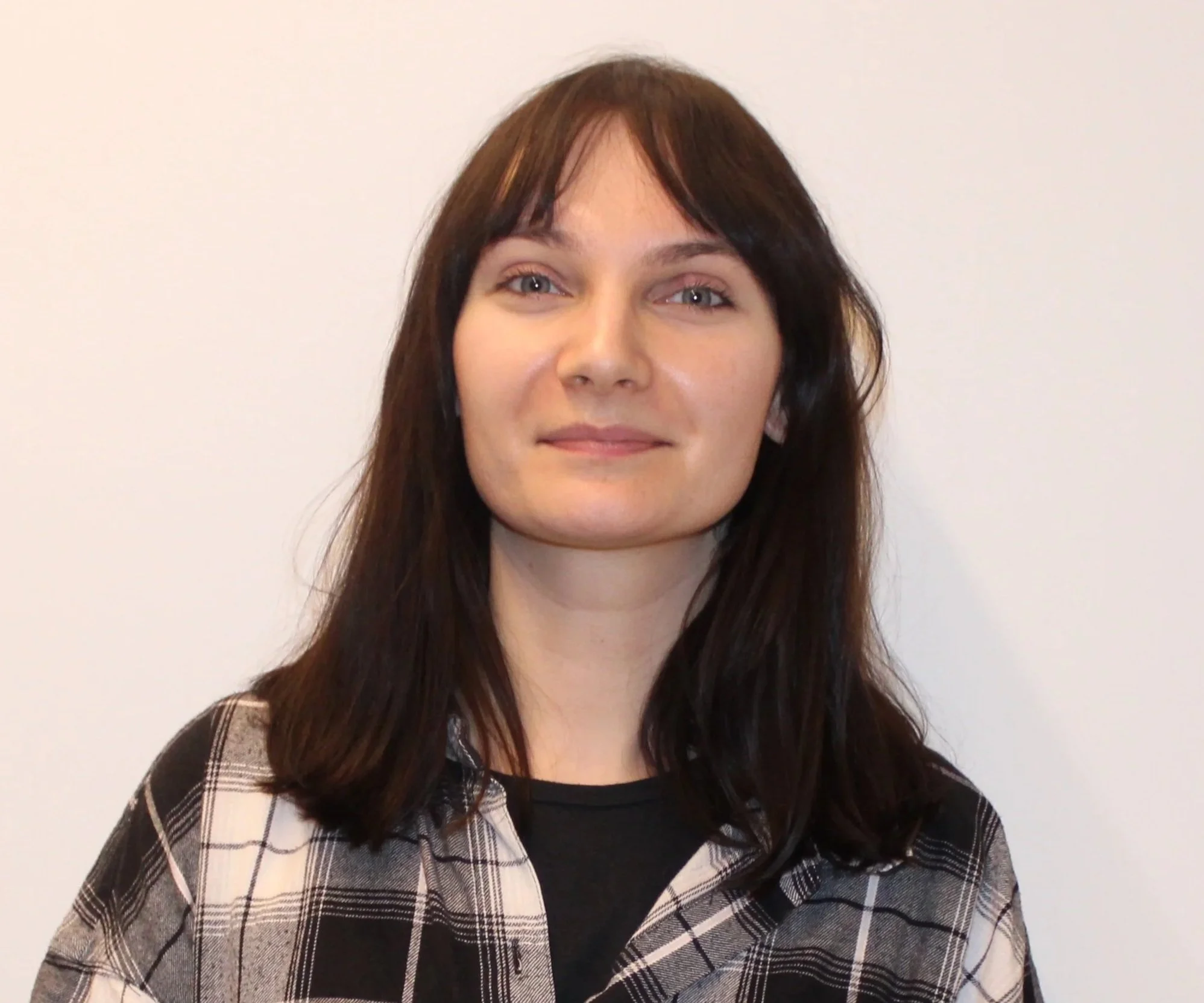 A young woman with shoulder-length dark brown hair and blue eyes, wearing a black shirt and a plaid button-up shirt, standing against a plain white wall.