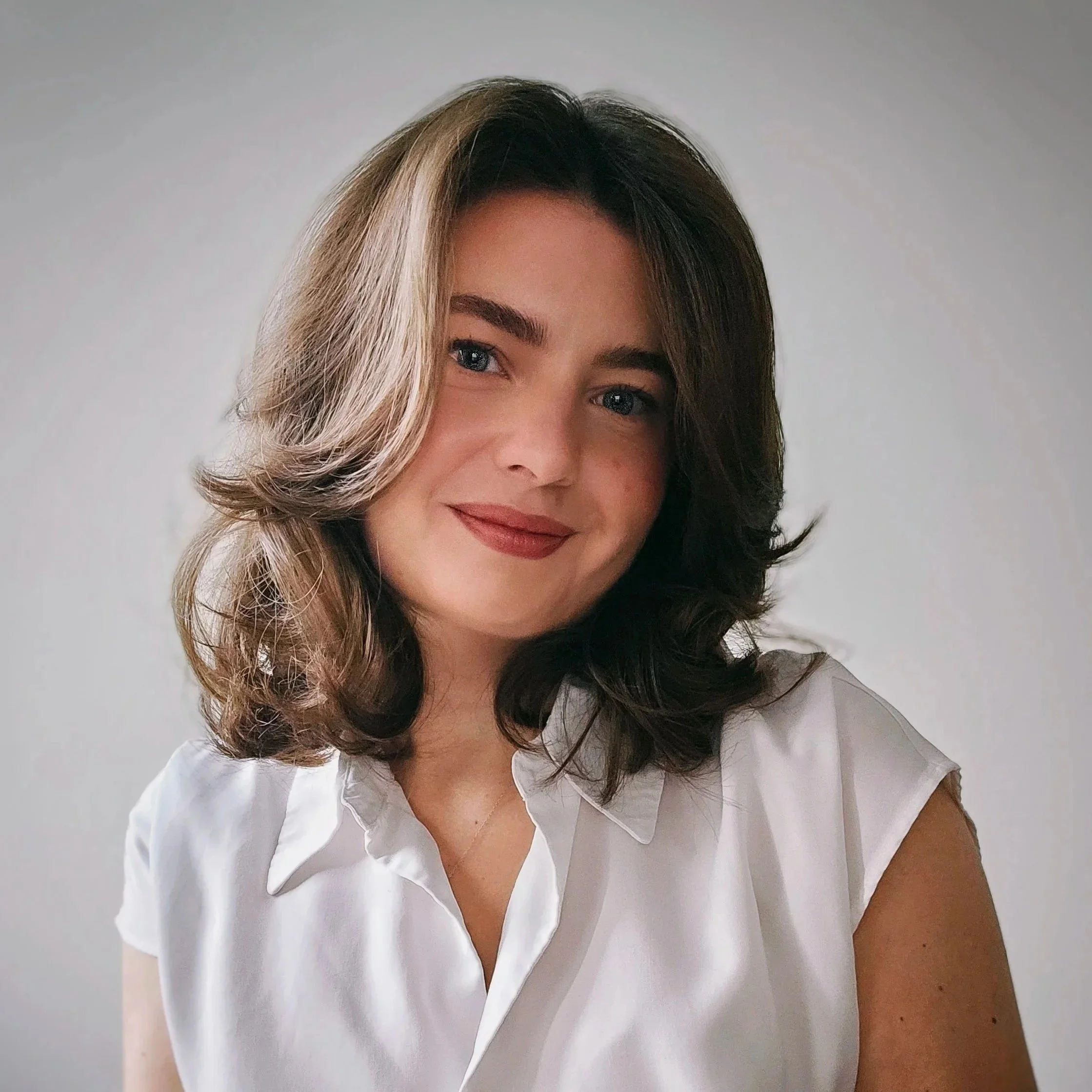 Portrait of a woman with shoulder-length dark hair wearing a light blue collared shirt and a gold necklace, smiling lightly against a plain white background.