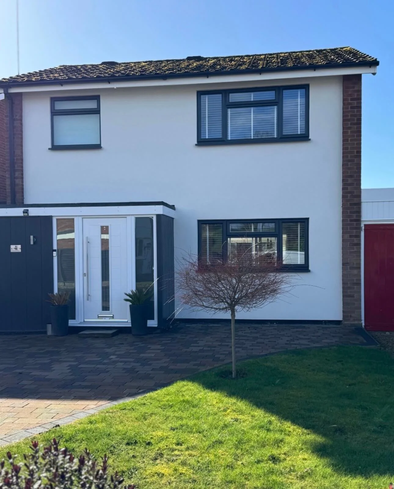 Front view of a two-story house with a white exterior, dark window frames, and a dark tiled roof. There is a small front yard with green grass and a young tree, along with a paved pathway leading to the front door, which is flanked by potted plants.