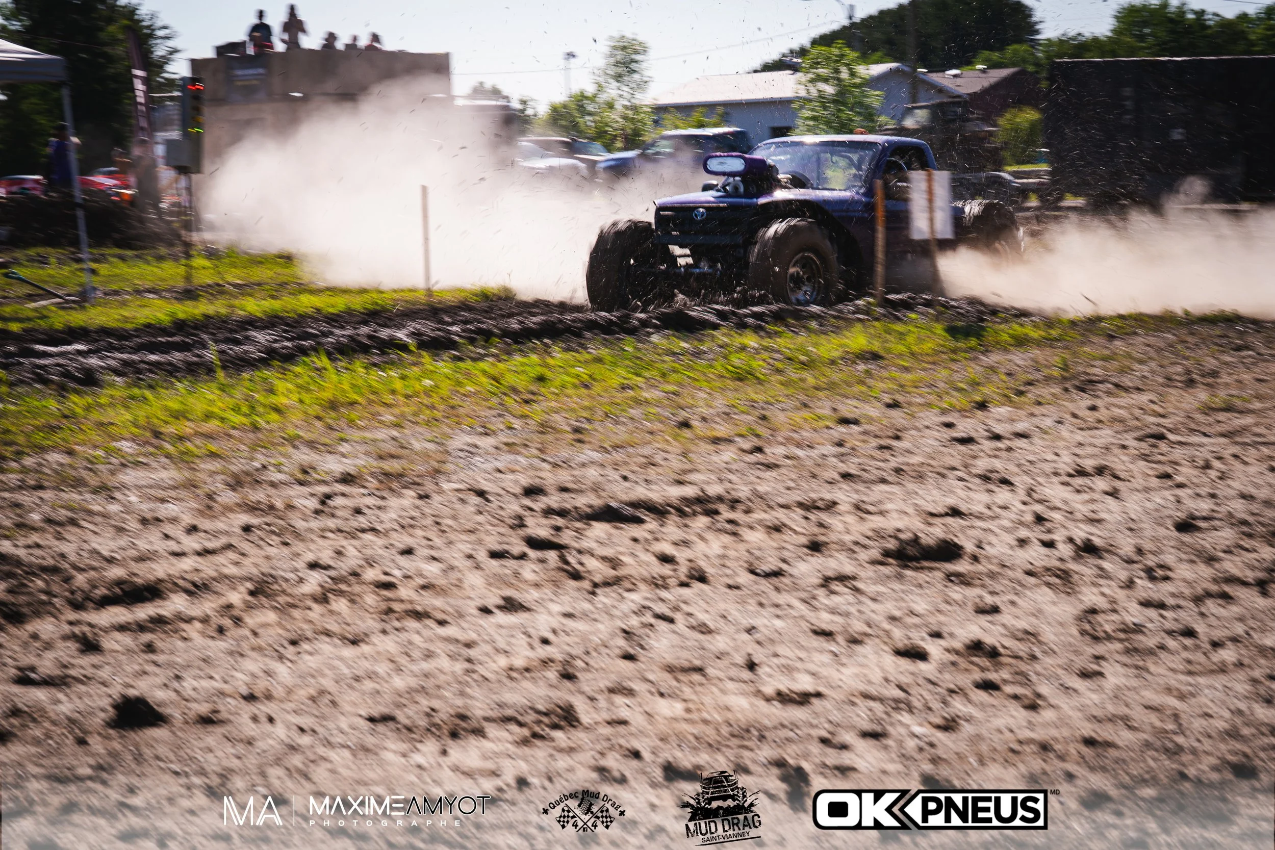 Un véhicule tout-terrain sur un parcours boueux lors d'une course de mud drag, avec poussière et fumée en arrière-plan, plusieurs spectateurs en haut et logos de sponsors au bas de l'image.