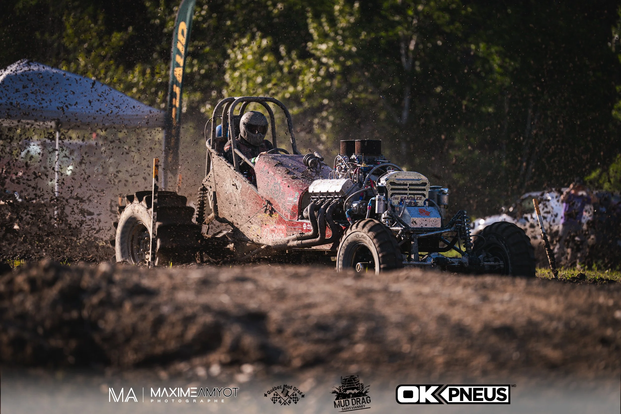 Voiture de mud racing traversant un terrain boueux avec un pilote portant un casque, entourée de poussière et de boue, sous un ciel ensoleillé.