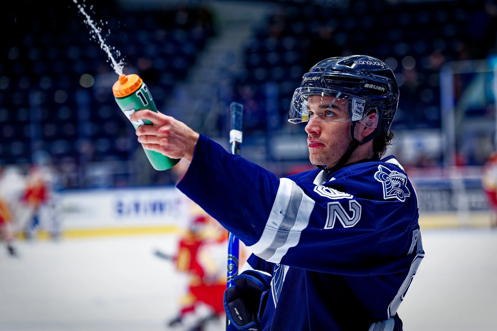 Ice hockey player in blue jersey and helmet spraying water from a Gatorade bottle on the ice during a game.