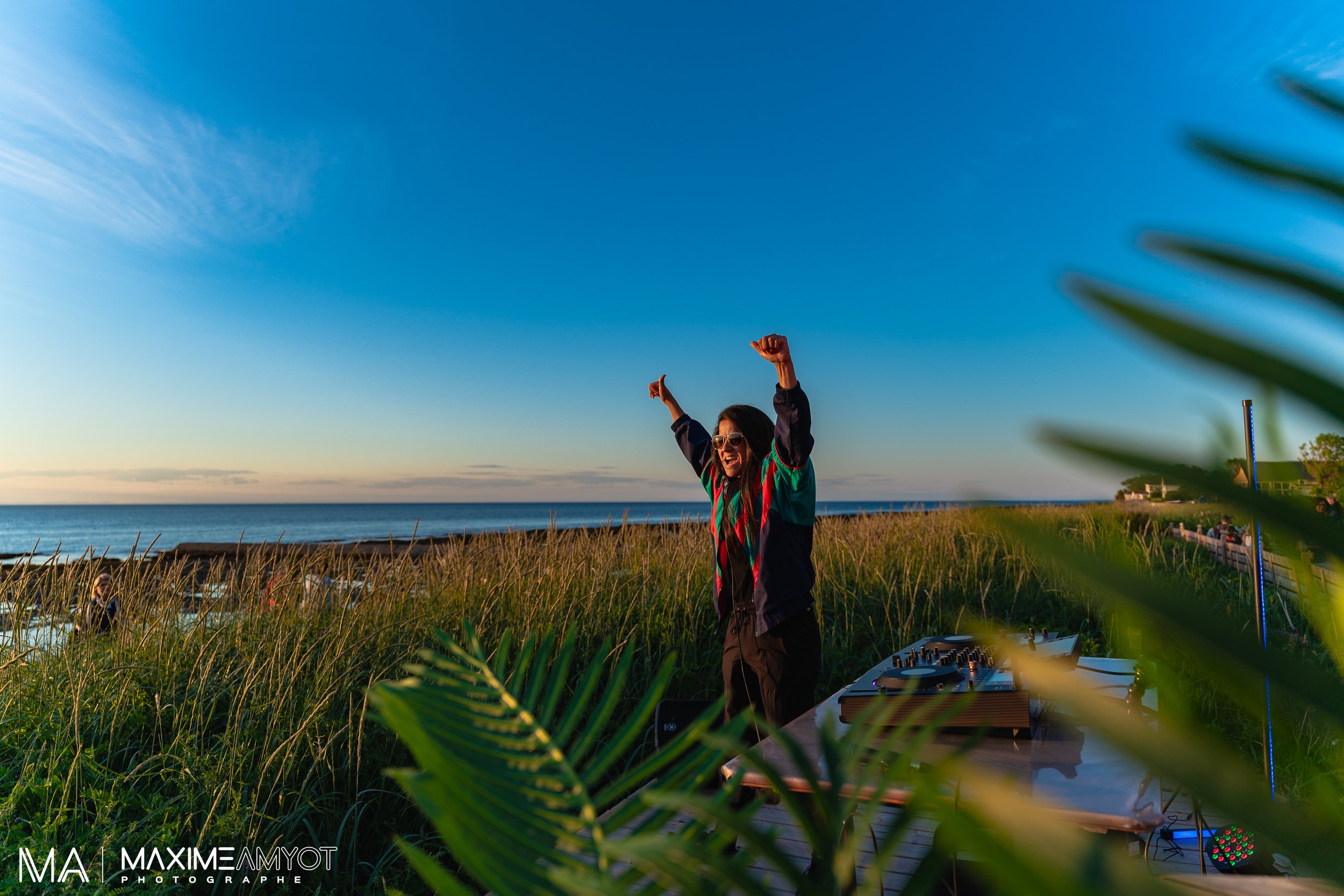 Une femme souriante avec des lunettes de soleil, levant les bras en l'air, semble danser ou célébrer en plein air près de la mer lors du coucher de soleil. Elle se trouve devant un équipement de DJ sur une table, entourée de hautes herbes et de végét