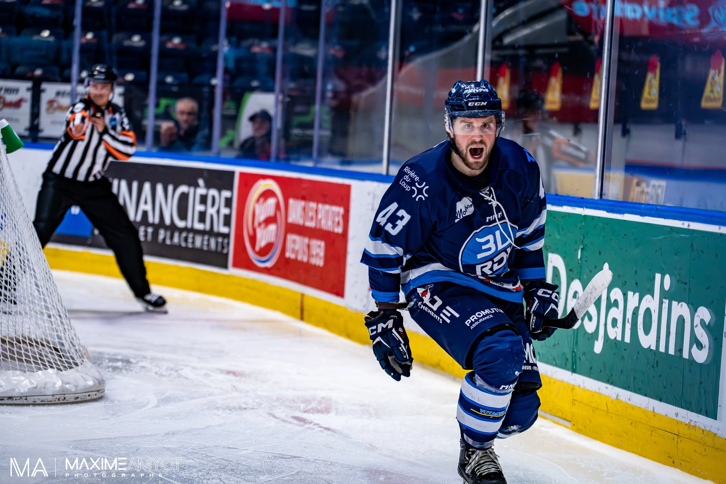 Un joueur de hockey en uniforme bleu, numéro 43, célèbre avec émotion sur la glace, avec un arbitre en arrière-plan et des spectateurs derrière la vitre.