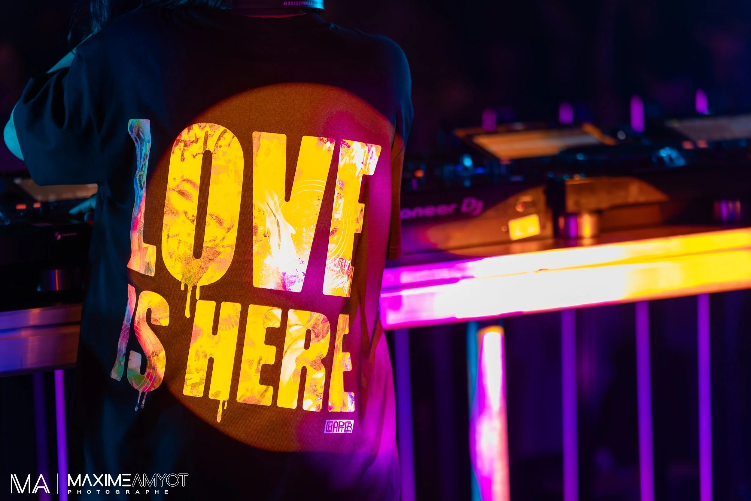 Un DJ jouant dans une boîte de nuit sombre, portant un t-shirt avec l'inscription "LOVE IS HERE" en lettres colorées Lumineuses