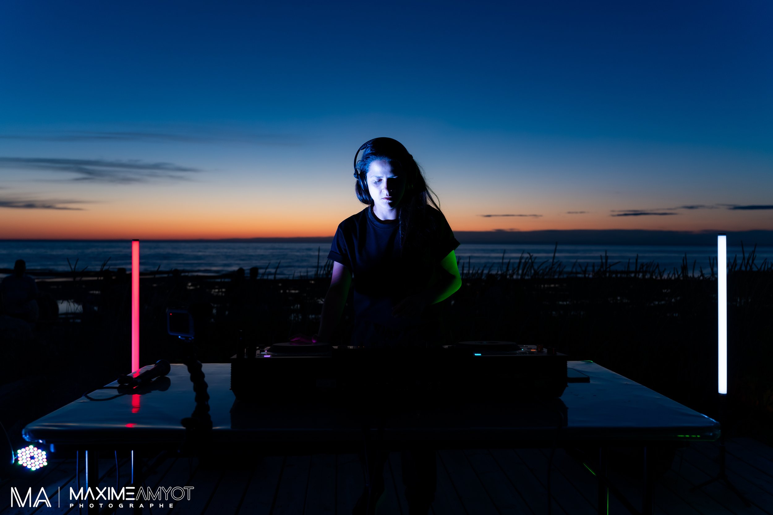 Une DJ joue de la musique lors d'un événement en plein air au coucher du soleil, avec vue sur la mer, éclairée par des lampes LED colorées.