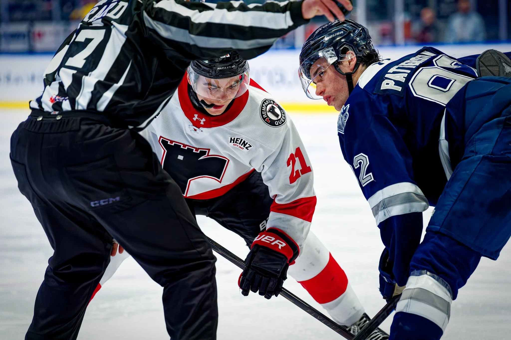 Hockey game face-off between two players and a referee on the ice. The player in a white jersey with red accents and the number 21 is facing off against a player in a blue jersey with the number 62. The referee is positioned between them, dropping th