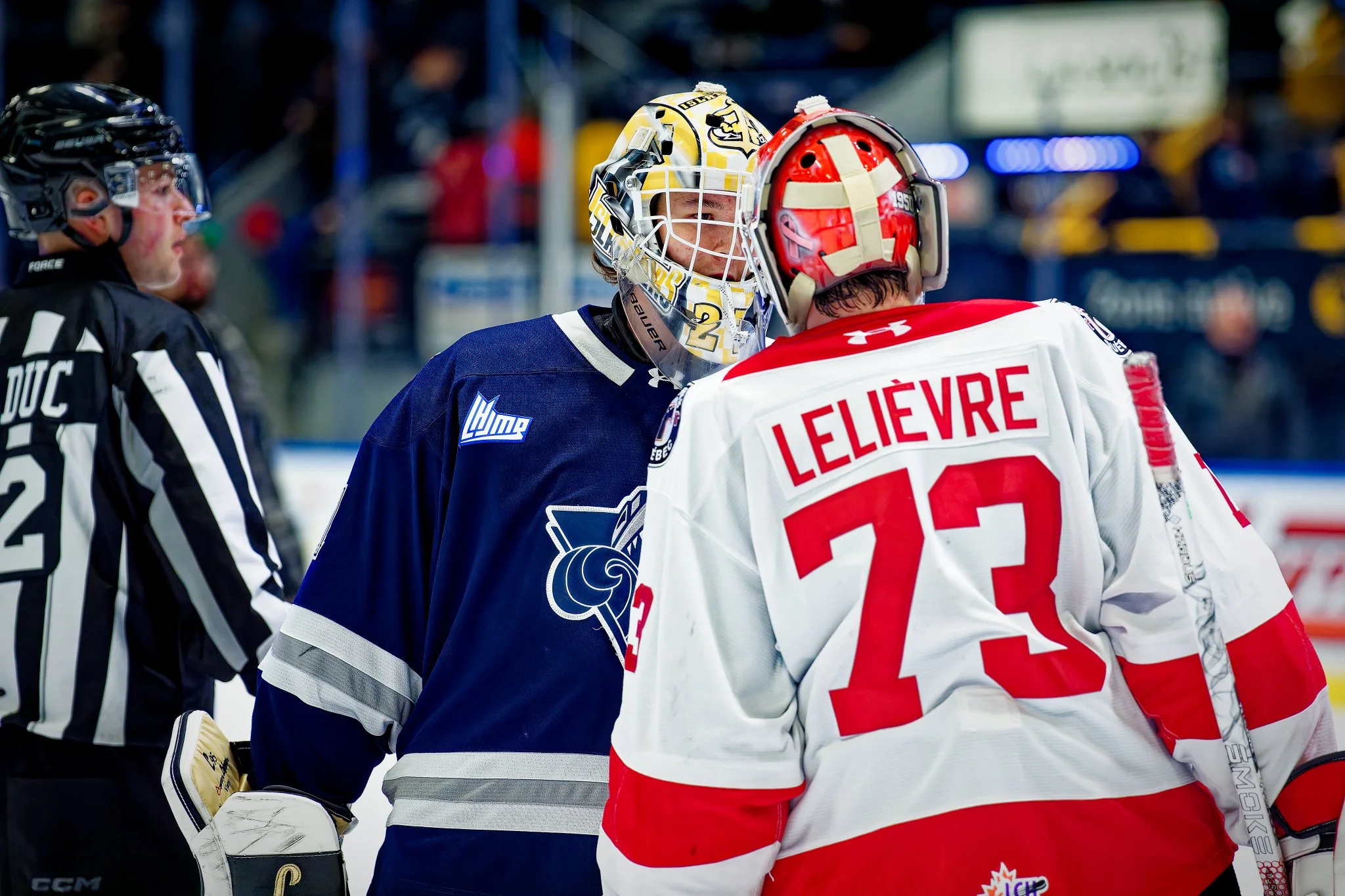 Ice hockey players and referee on the ice rink, two players facing each other, one in a navy blue jersey and the other in a white and red jersey, with a stadium in the background.