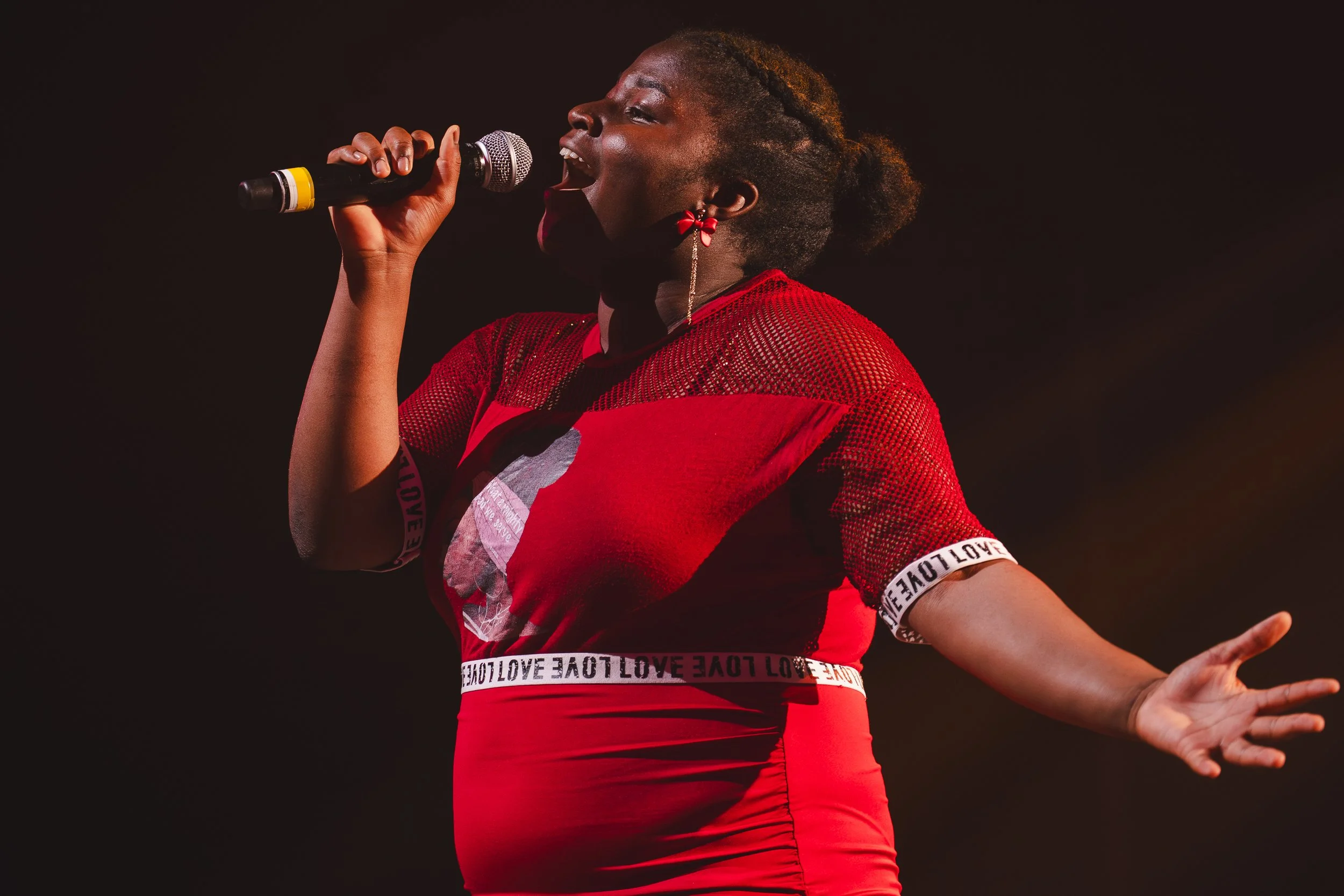 Une femme chante en portant un vêtement rouge avec des détails noirs, tenant un microphone sur scène, avec une expression passionnée.