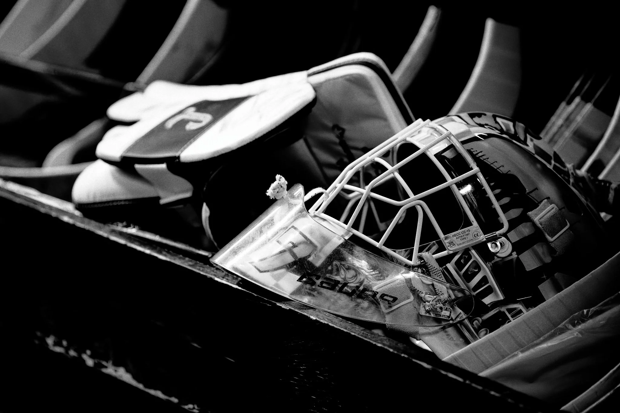 Black and white photograph of a hockey goalie mask and a glove placed on a hockey bench.