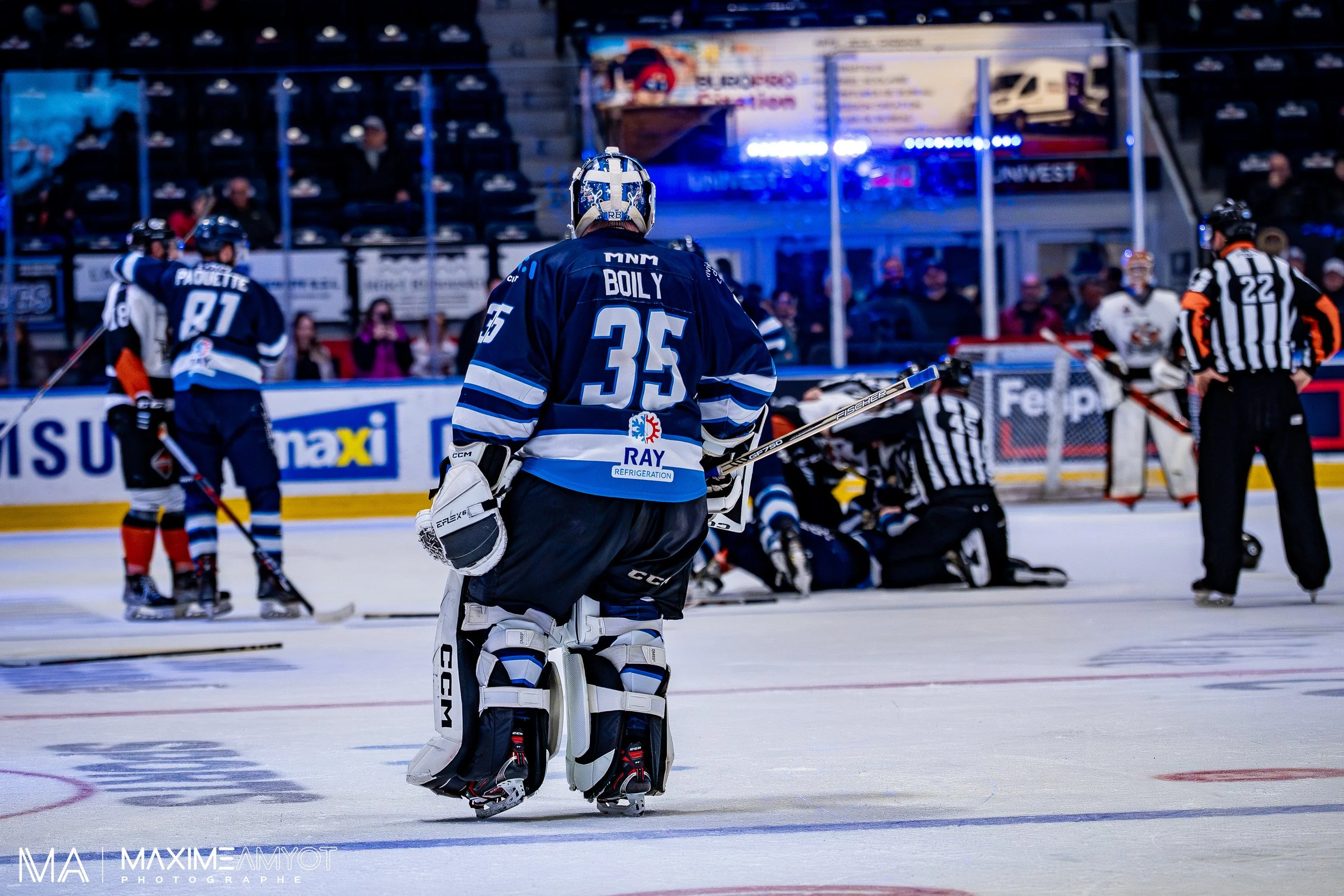Joueur de hockey sur glace portant le numéro 35, debout sur la glace lors d'un match, avec d'autres joueurs, un arbitre et un infirmier en arrière-plan.