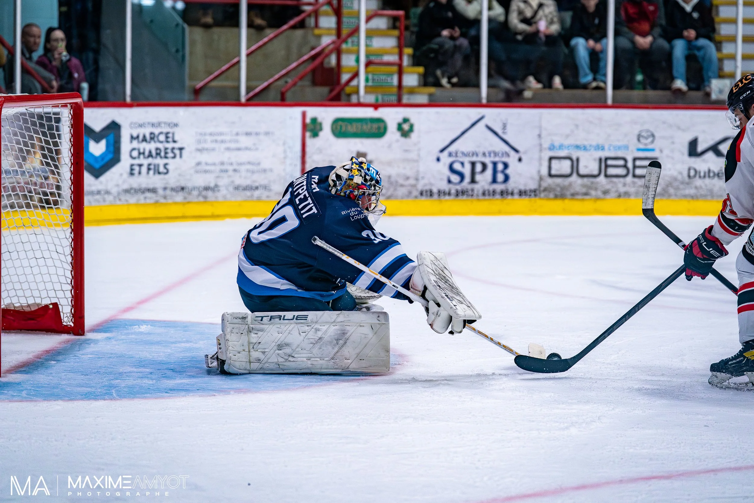 Un gardien de but de hockey sur glace en position de plongeon devant la cage, tentant de bloquer un palet que l'adversaire tente de tirer dans le but. L'adversaire porte un maillot blanc avec des rayures rouges et noires. Des spectateurs sont visible