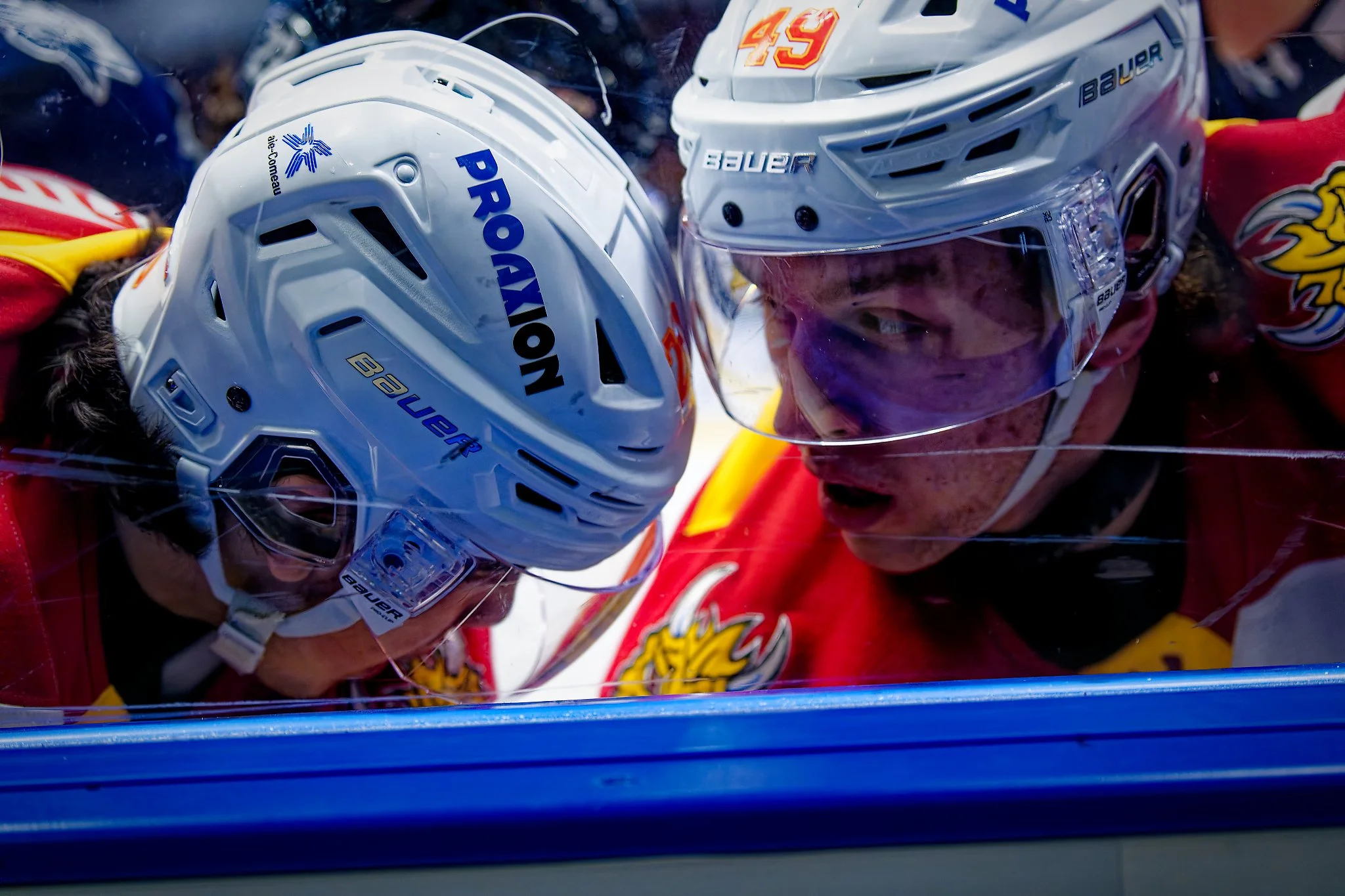 Two hockey players in helmets and jerseys engaged in a close interaction, with one player wearing a white helmet and the other a white helmet with a yellow and red logo, both wearing maroon jerseys with yellow accents.
