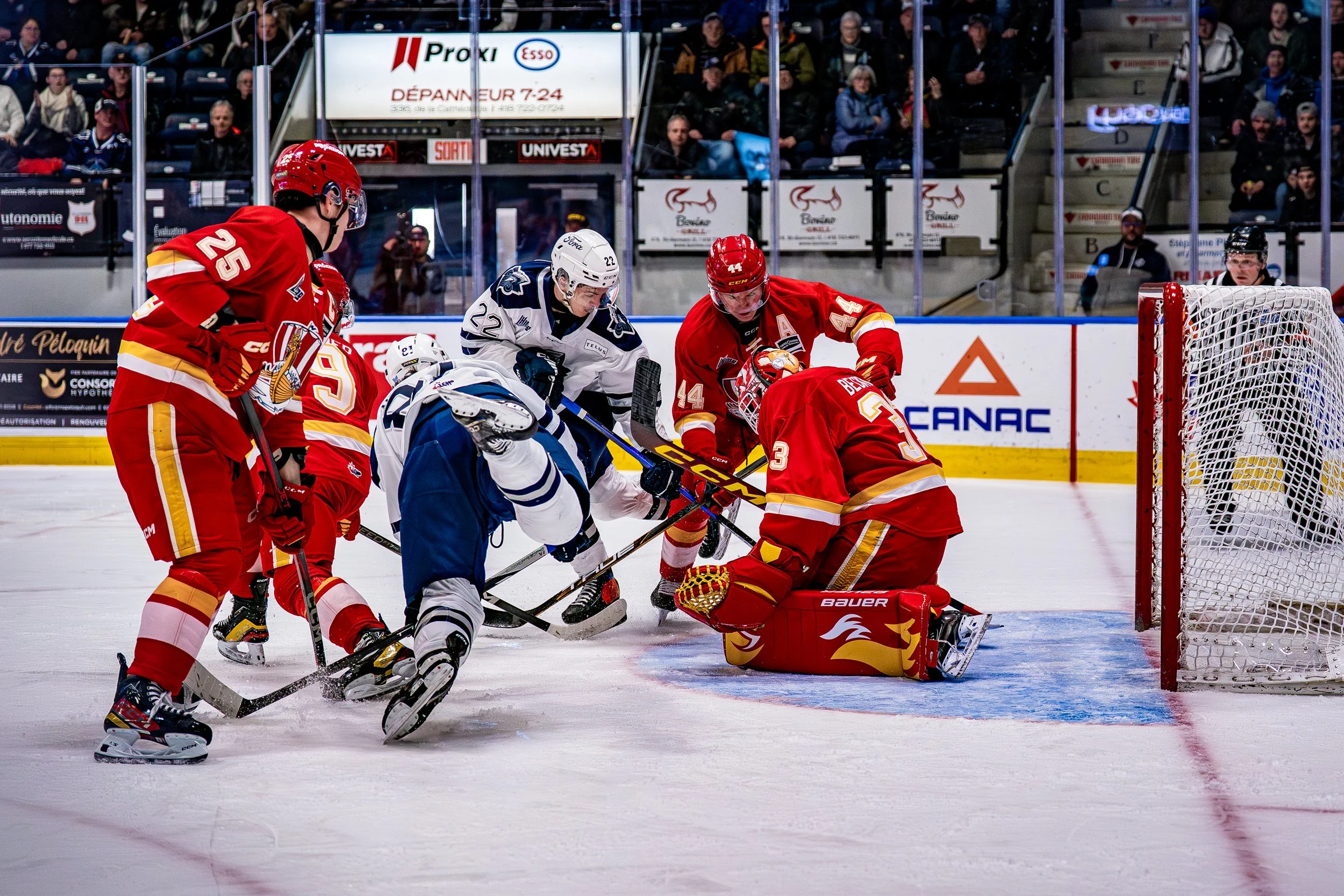 Joueurs de hockey sur glace en action, un joueur en maillot blanc tente de marquer devant le but gardé par un gardien en équipement rouge, avec d'autres joueurs en maillots rouges et blancs autour dans une patinoire, spectateurs derrière.