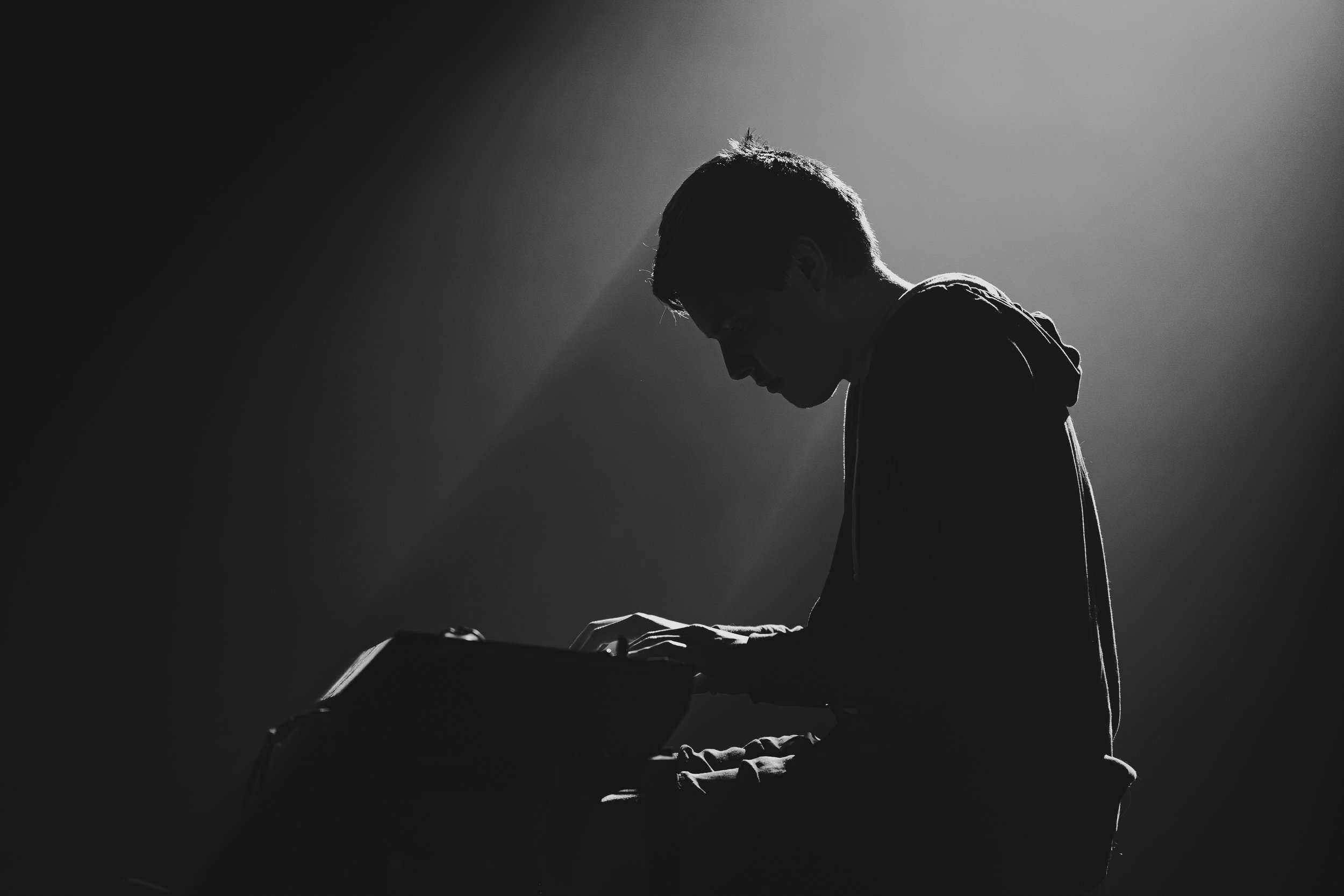 Silhouette d'un jeune homme jouant du clavier sur scène, éclairé par une lumière douce. Environnement sombre, ambiance de musique ou de performance artistique.