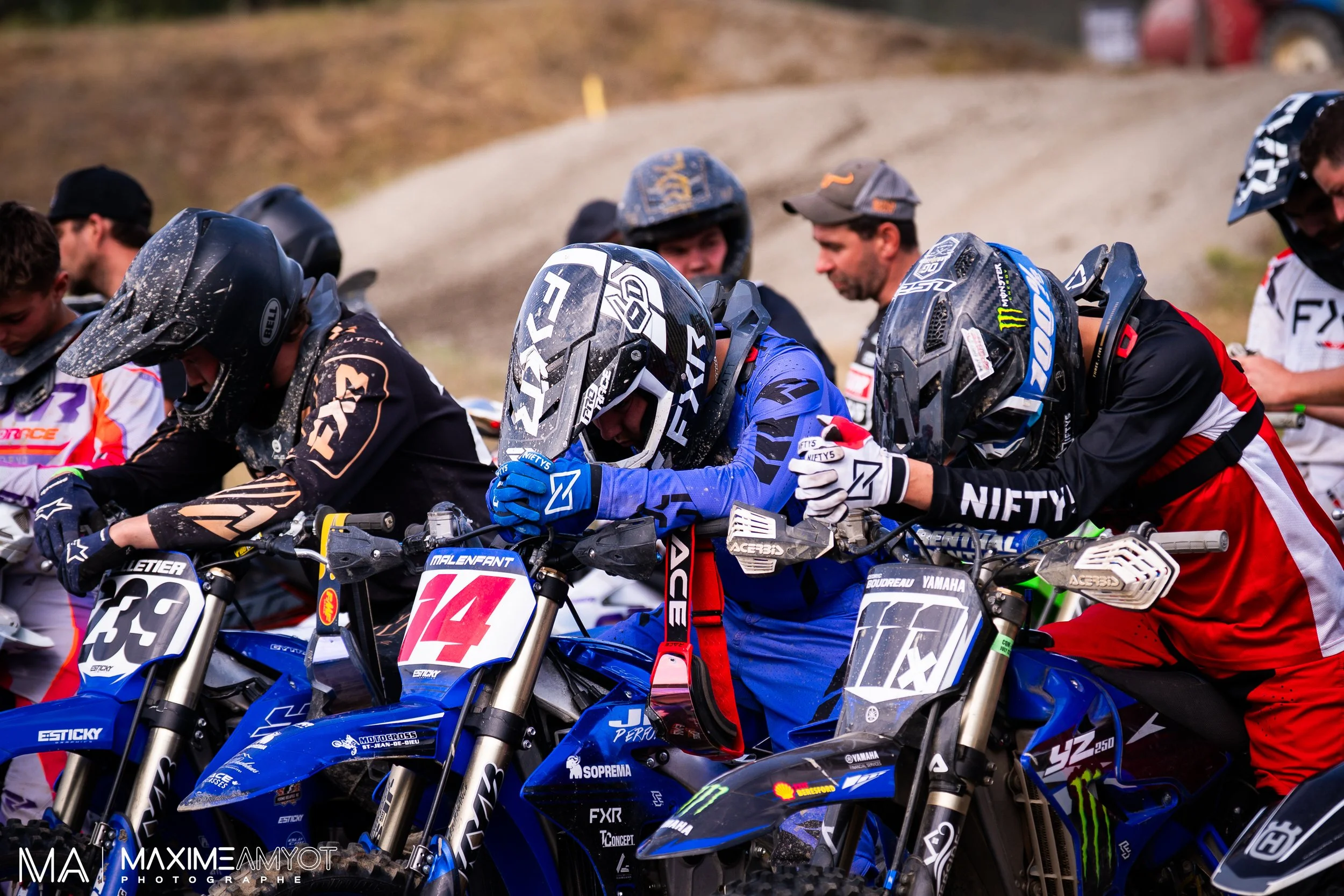 Group of motocross racers in gear and helmets preparing for a race, standing beside their bikes on a dirt track.