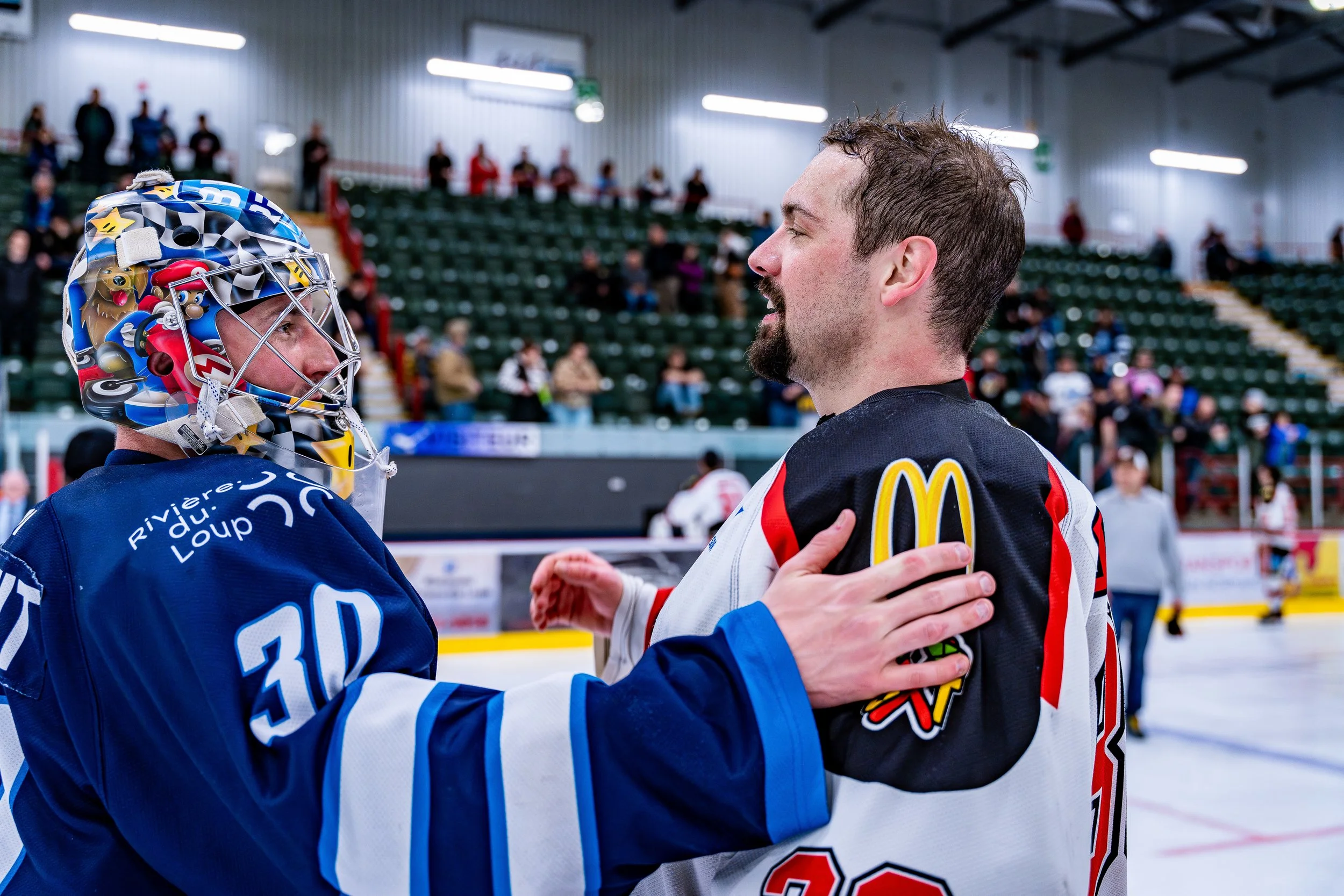 Deux joueurs de hockey s'encadrent et se saluent après un match sur la glace, en présence de spectateurs dans les gradins.