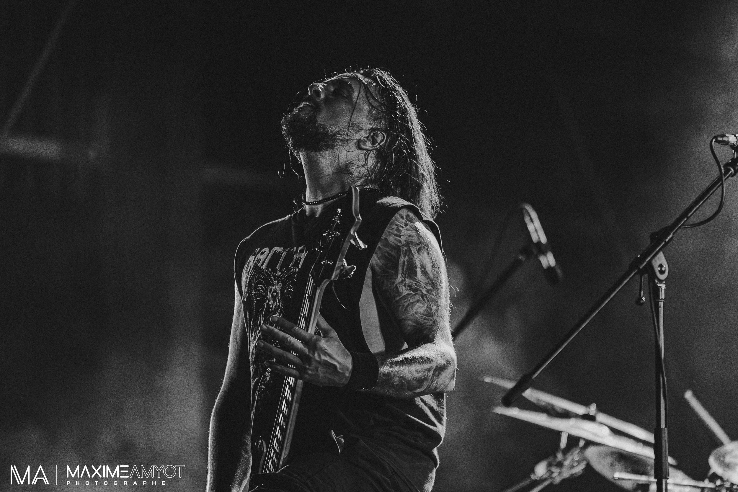Musicien de rock avec tatouages, cheveux longs, et barbe, jouant de la guitare sur scène en noir et blanc, avec microphones et cymbales en arrière-plan.