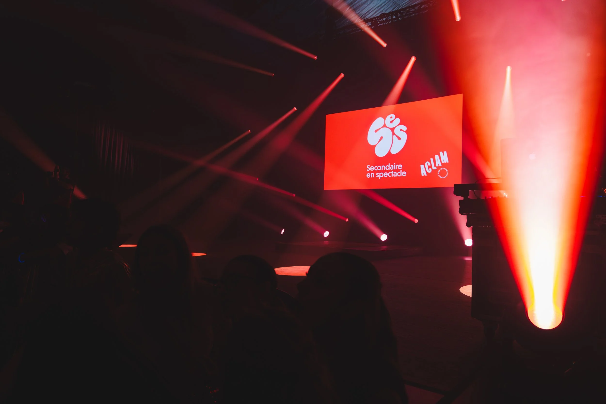 Scène de spectacle avec un écran rouge affichant un logo, le texte 'Secondaire en spectacle' et 'ACLAM', entourée de lumières et de fumée. Silhouettes de personnes dans l’obscurité au premier plan.