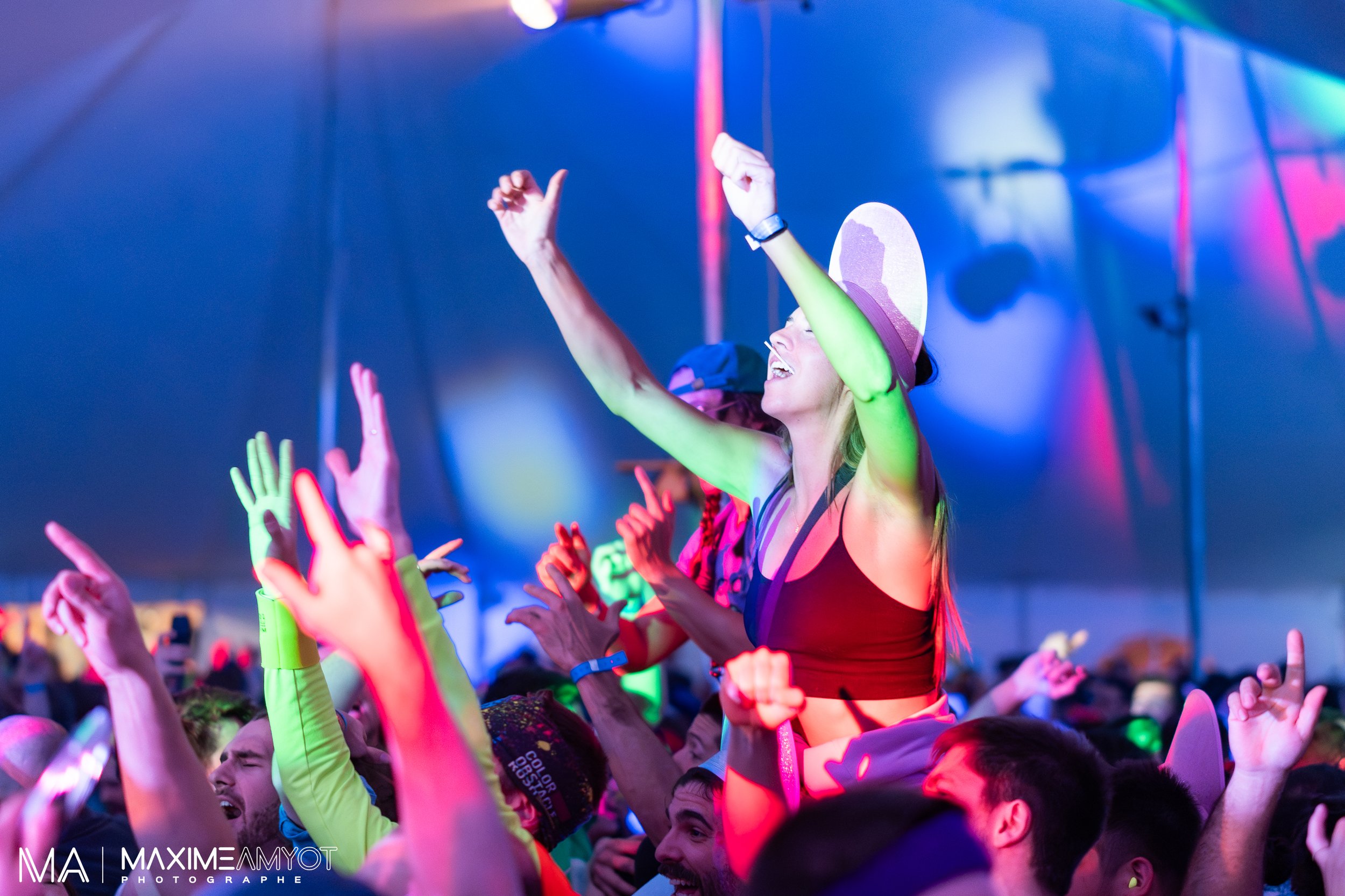 Groupe de personnes dansant dans un festival ou concert, sous un chapiteau, avec des lumières colorées et une femme portant un chapeau et souriant.