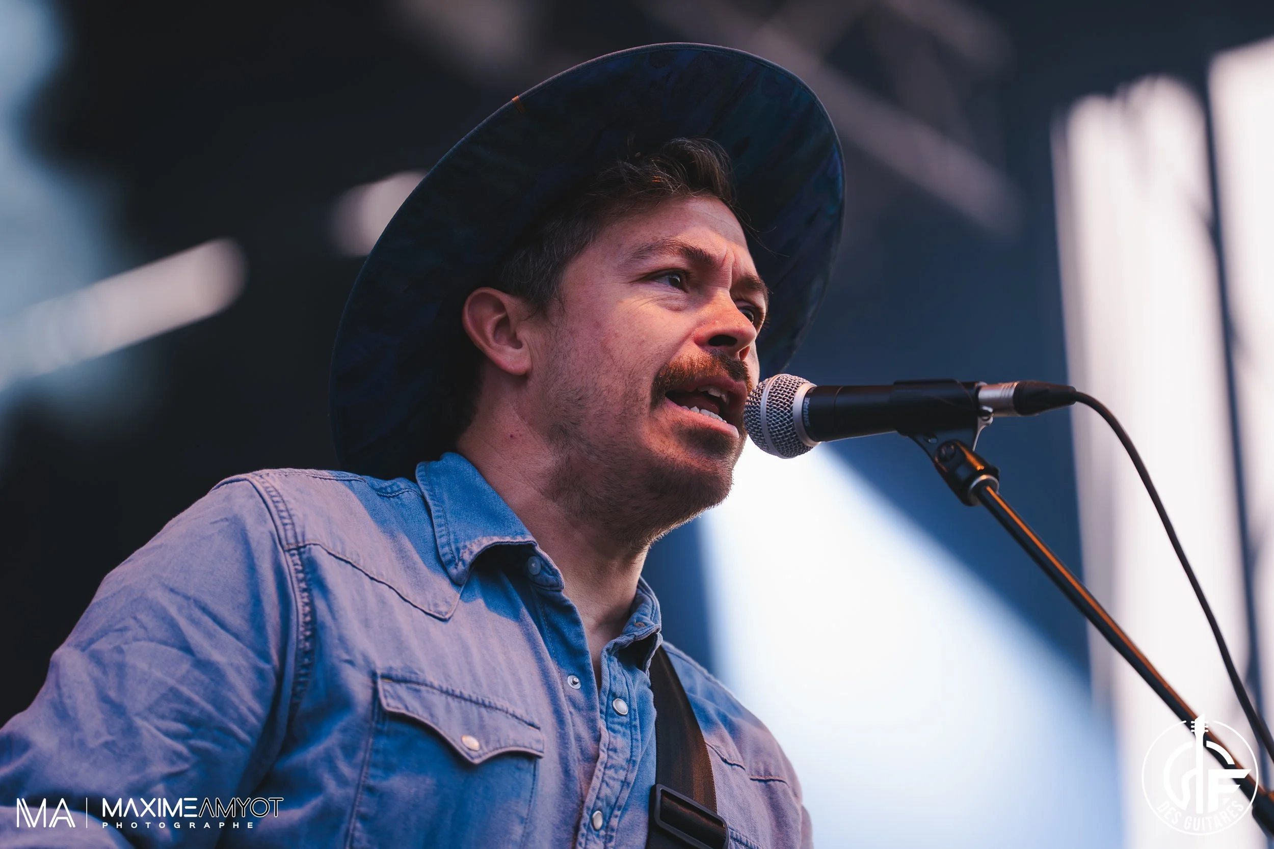Homme chantant ou parlant dans un microphone lors d'une performance, portant un chapeau large et une chemise en denim.
