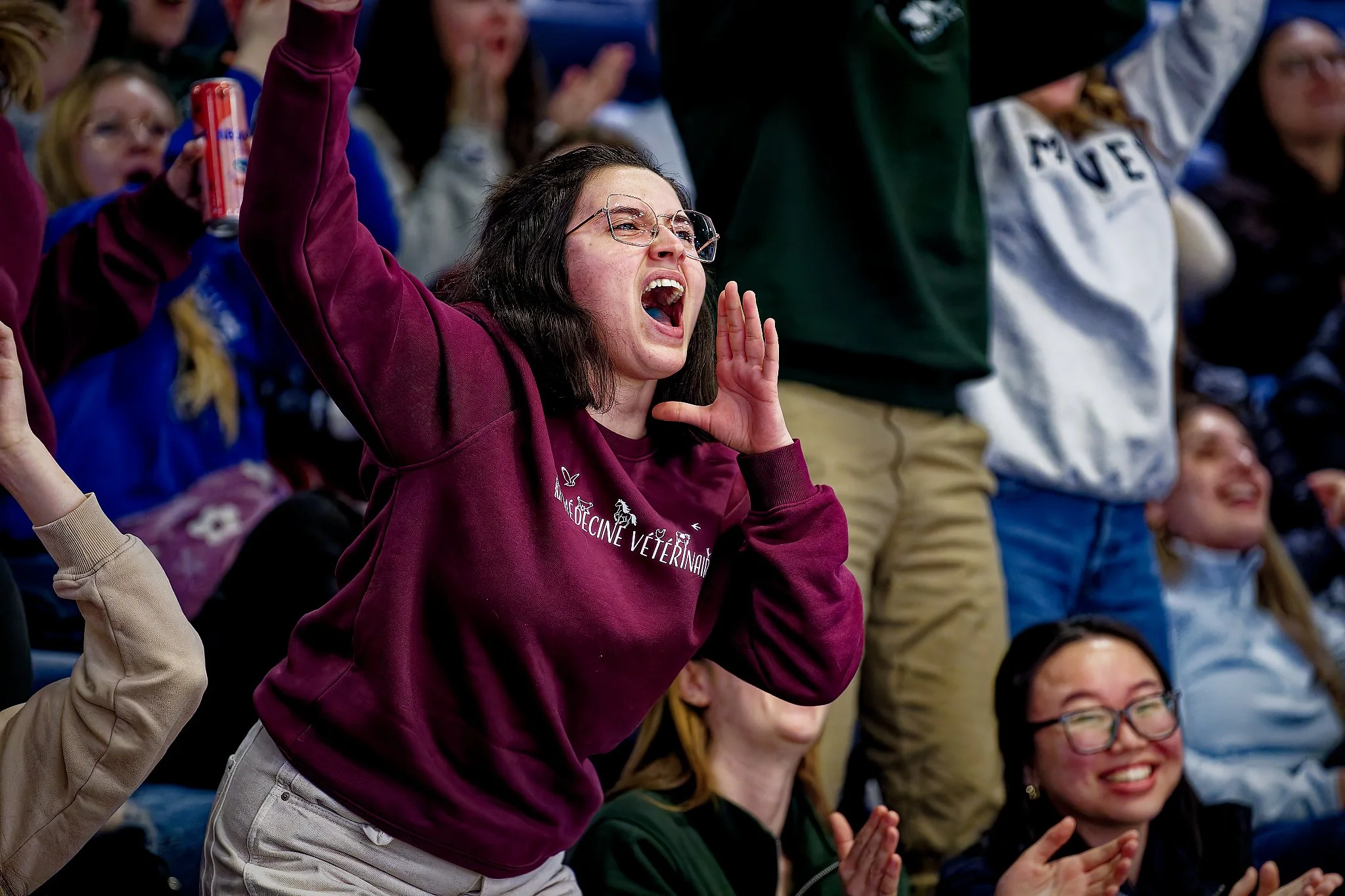 A woman cheering enthusiastically at a sports event, wearing glasses and a maroon sweatshirt that says 'VETERINARY' on it, surrounded by other people also cheering and clapping.