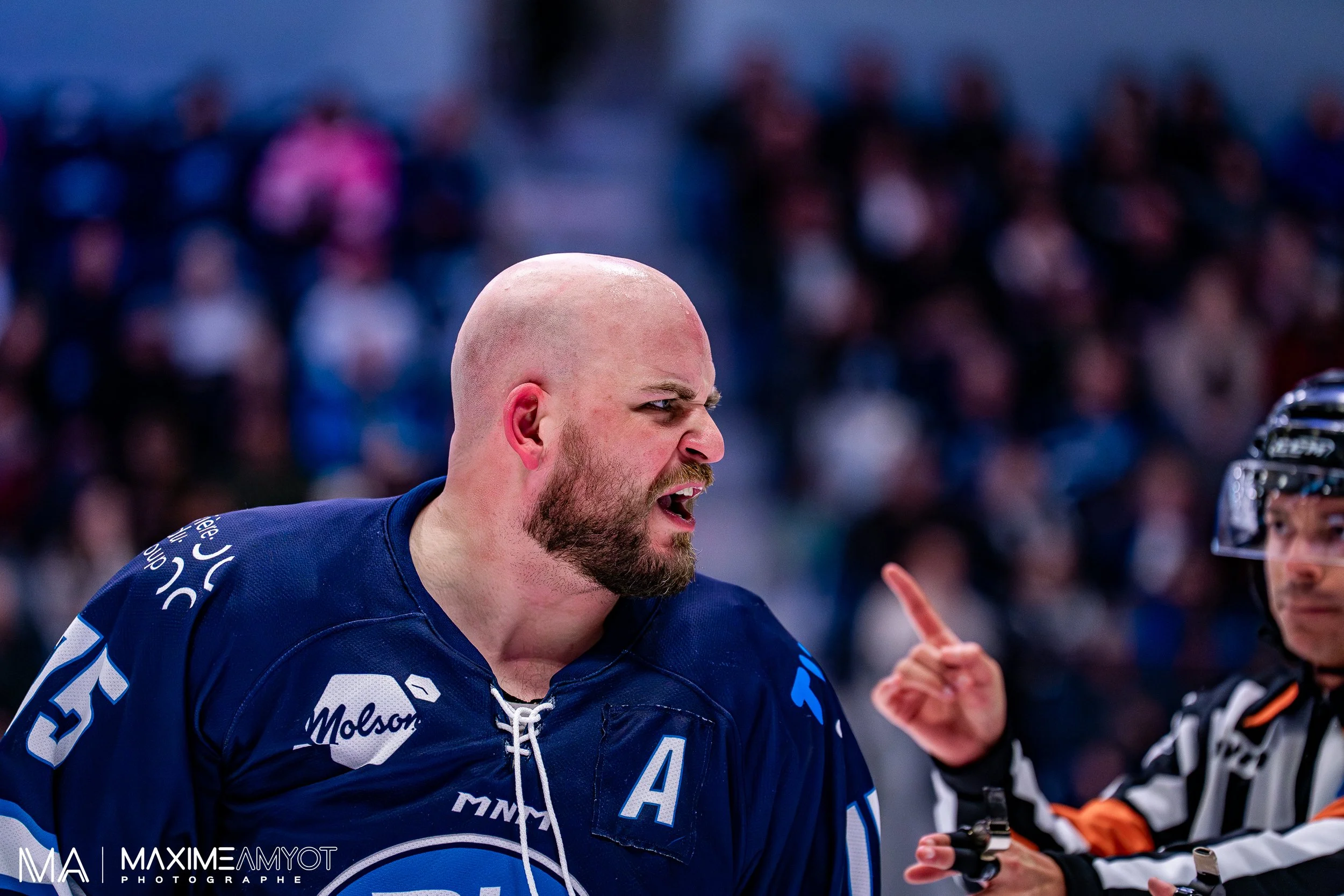 Un joueur de hockey en colère, gesticulant vers un arbitre lors d'un match, portant un maillot bleu avec un symbole 'A' sur la poitrine, dans une enceinte sportive entourée de spectateurs.
