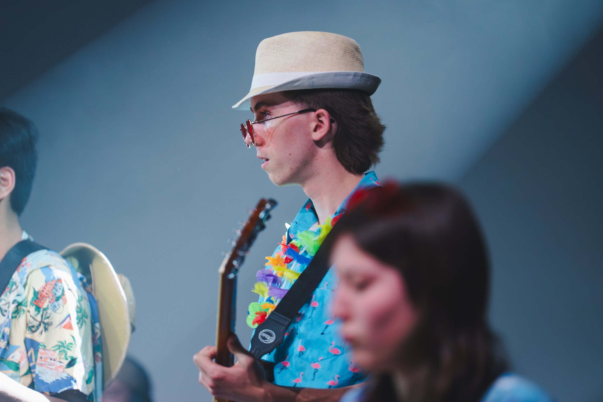 Un jeune homme avec des lunettes de soleil colorées, portant un chapeau de paille et une chemise hawaïenne à motif flamants roses, joue de la guitare lors d'une réunion ou d'un événement.