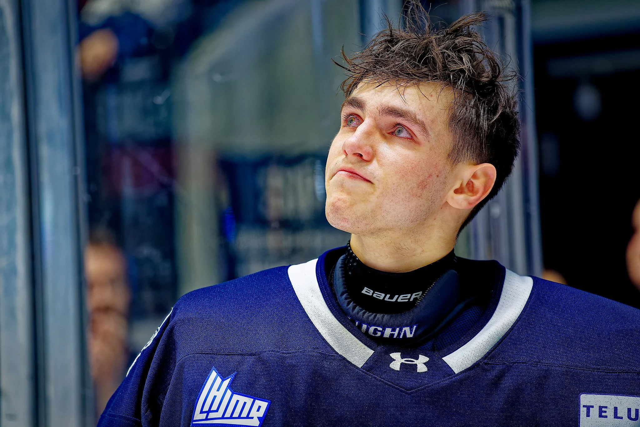 Close-up of a young male ice hockey player with short brown hair, tears in his eyes, wearing a navy blue jersey with the team logo and a black neck protector, standing near the ice rink.