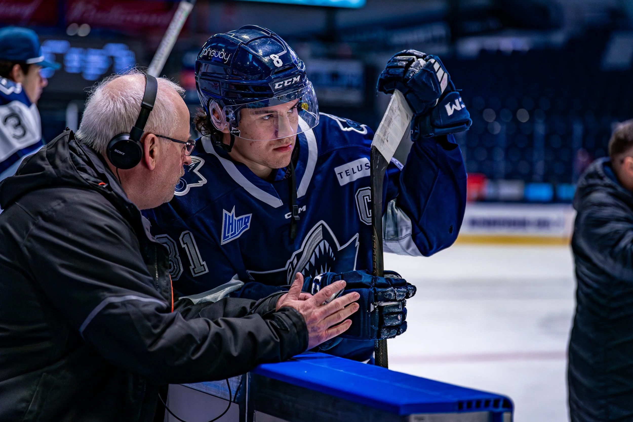 Un joueur de hockey en uniforme bleu écoutant un entraîneur avec un casque audio pendant une séance sur la glace.