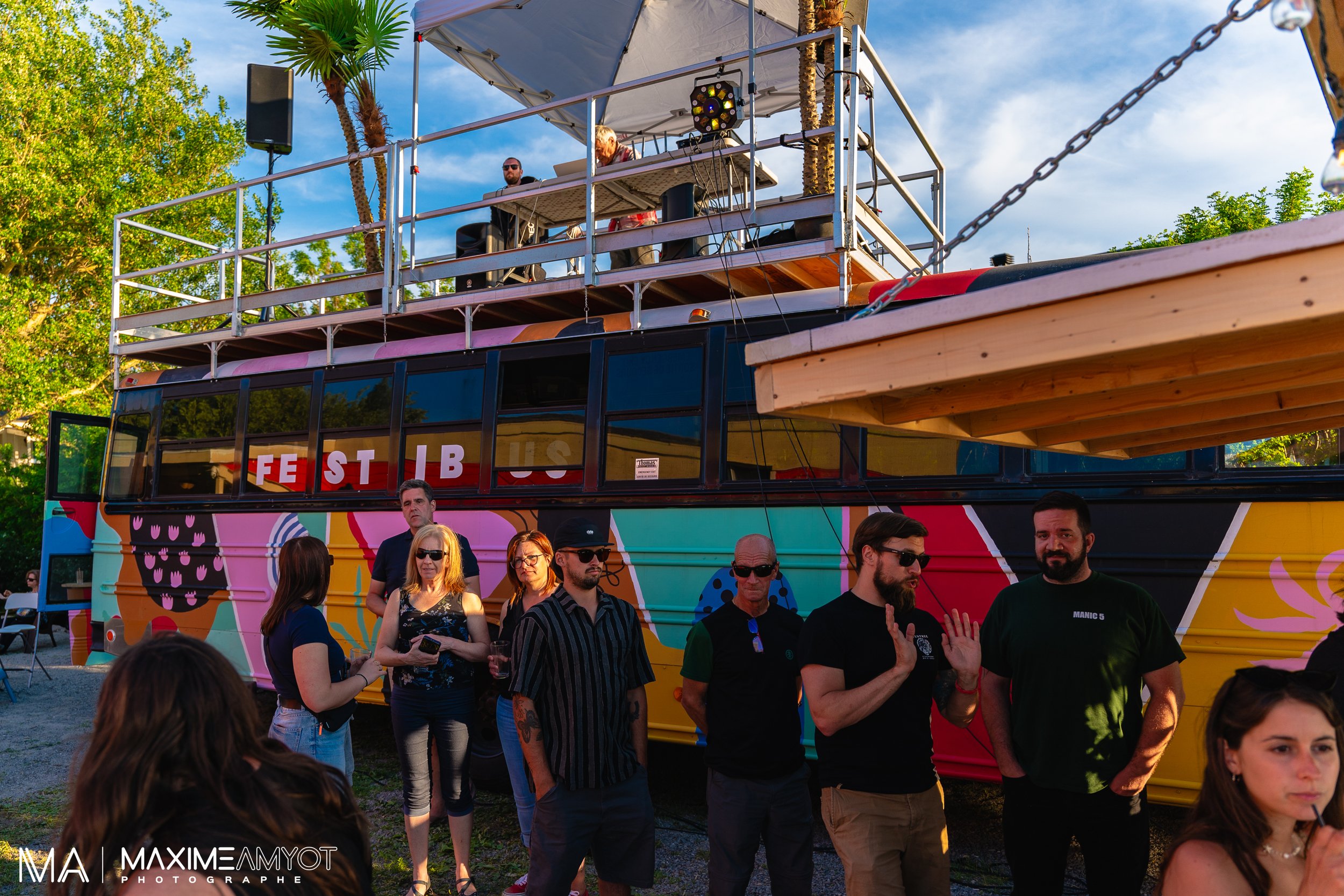 Un groupe de personnes se tient devant un bus coloré lors d'un festival en plein air, avec une scène sur le toit du bus où deux DJ jouent de la musique. Le bus a une peinture vibrante avec des motifs graphiques et le mot 'FESTIBUS' écrit dessus, sous