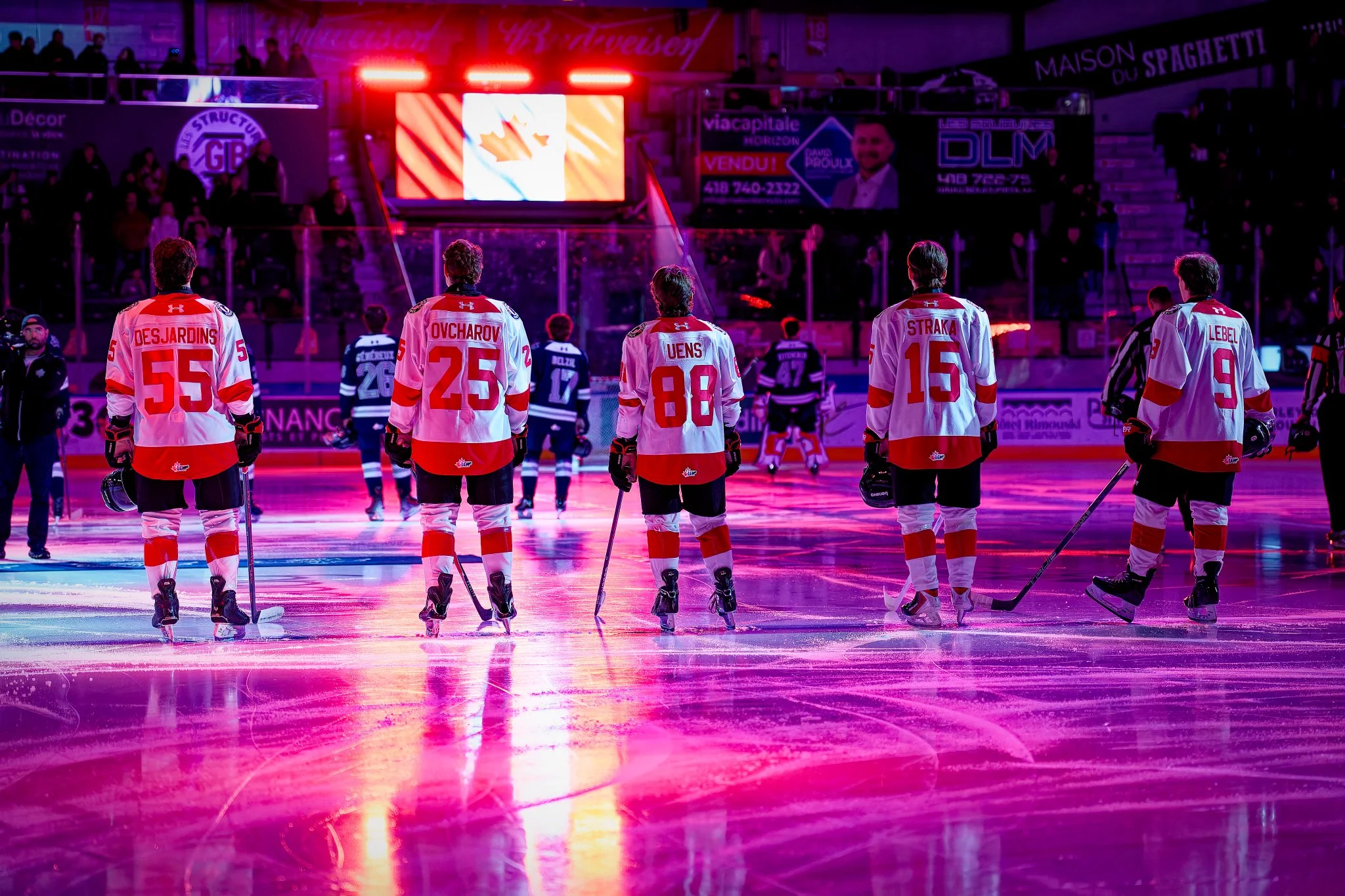 Ice hockey players in white and red jerseys standing on the ice rink, facing the scoreboard, with purple lighting and spectators in the background.