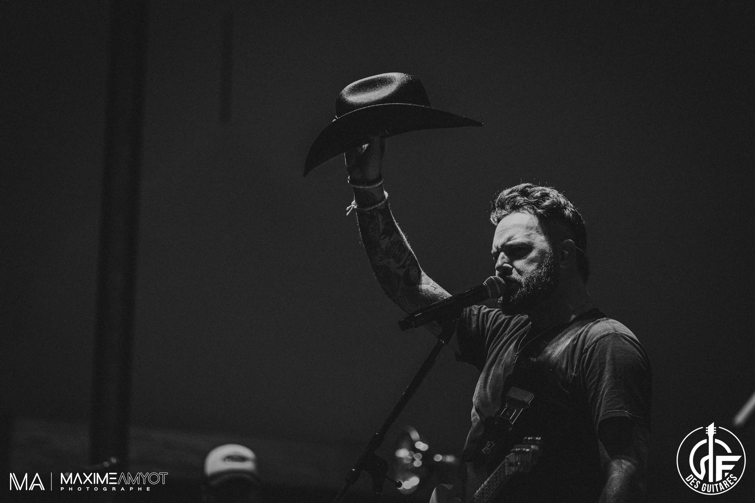 Musicien sur scène en noir et blanc, levant son chapeau, chantant dans un microphone, avec une guitare en bandoulière.