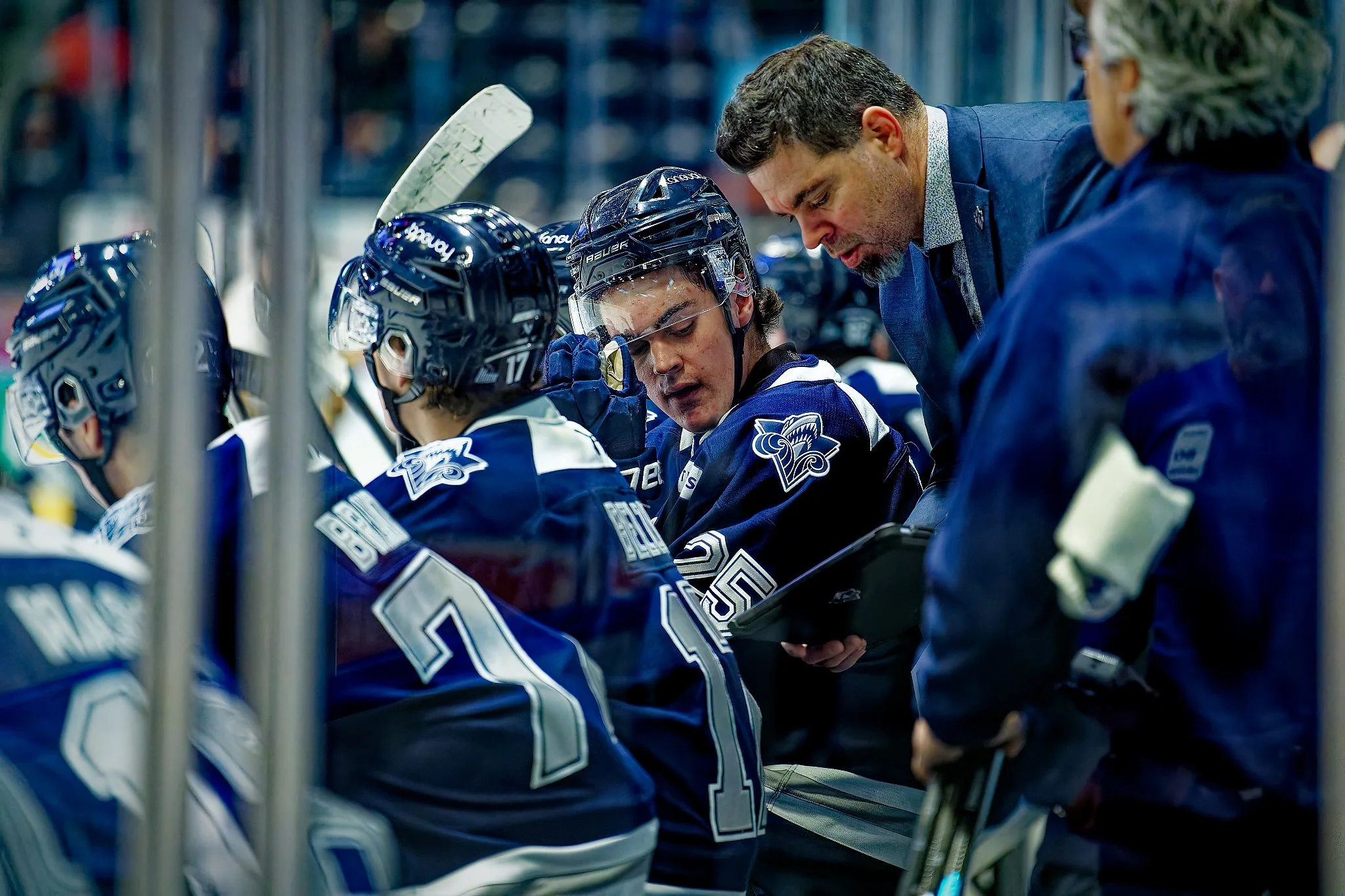 Hockey players on the bench during a game, wearing blue jerseys and helmets, with coaches giving instructions.