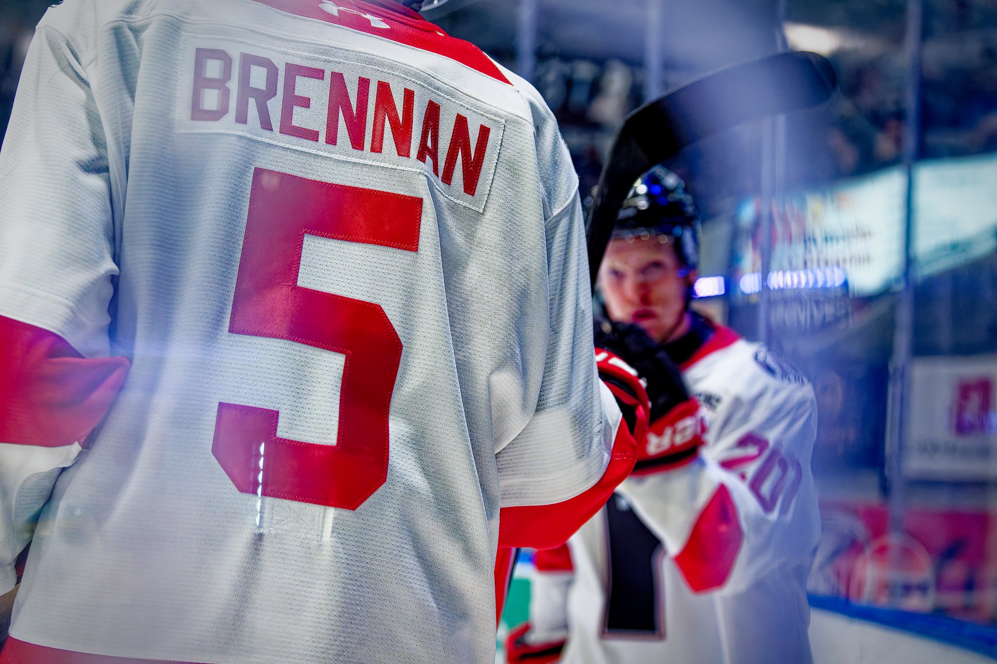 Close-up of an ice hockey player’s jersey with the name "Brennan" and the number 5, in an ice rink with another player in the background.