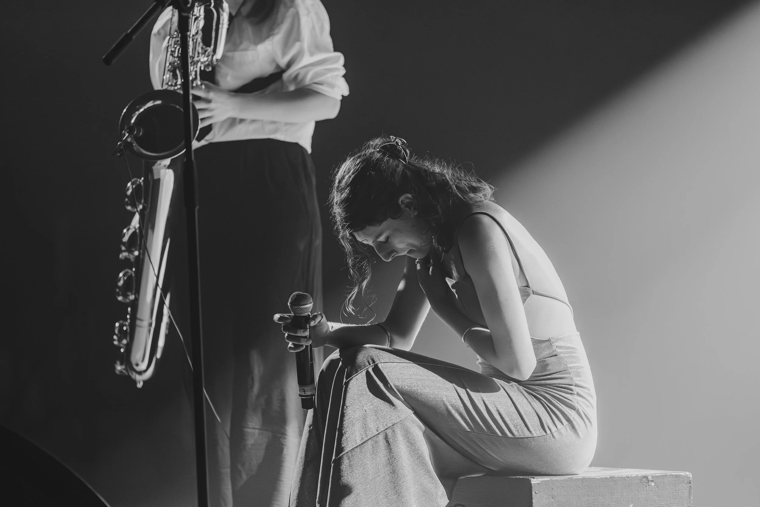 Une femme assise sur un banc, tenant un micro, en pleurs ou profondément émue, lors d'une performance ou d'une scène artistique, avec un musicien jouant de la saxophone en arrière-plan dans une ambiance sombre et dramatique.
