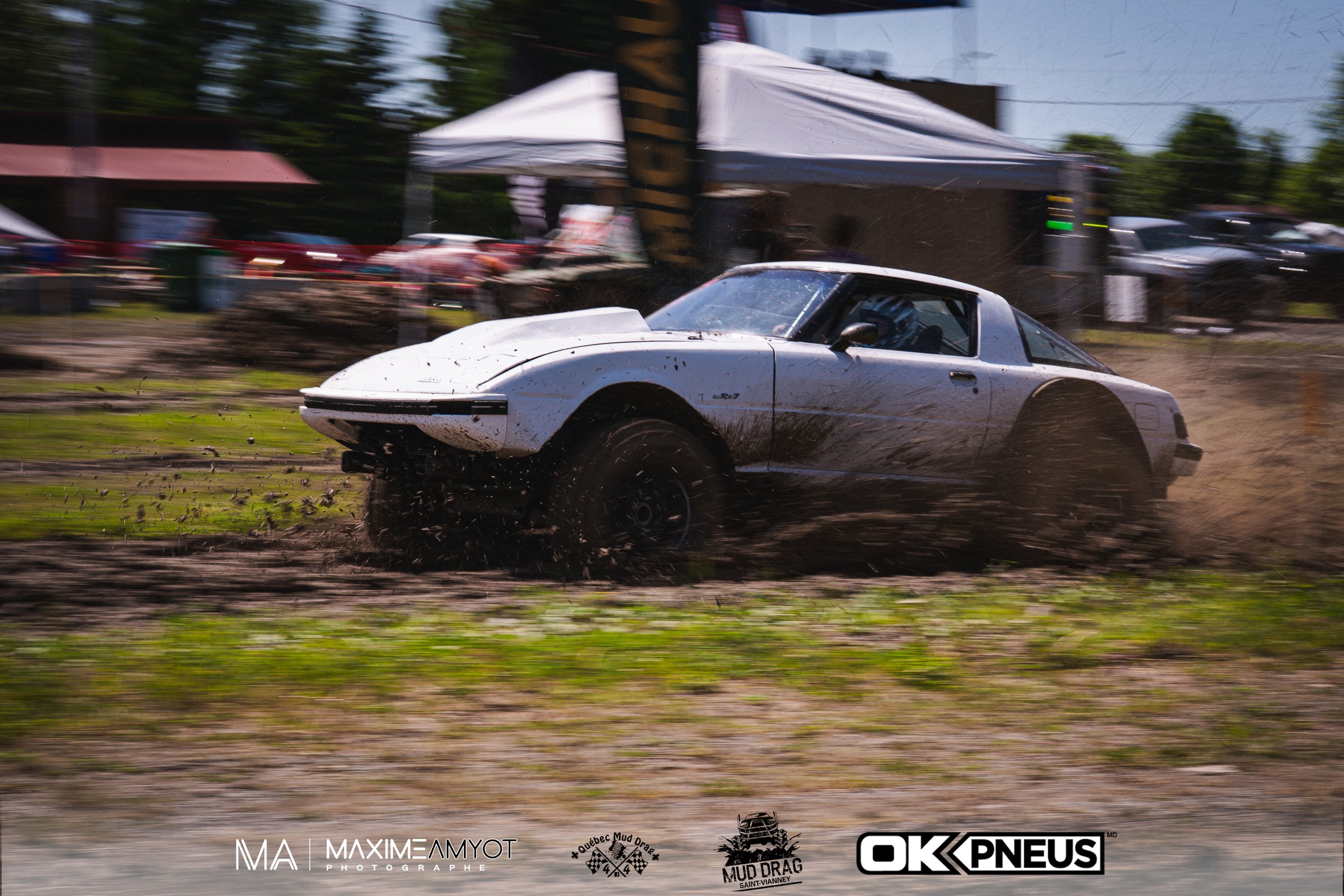 Une voiture de course blanche dérape en parcourant un terrain boueux lors d'une compétition de Mud Drag avec des tentes et des spectateurs en arrière-plan.