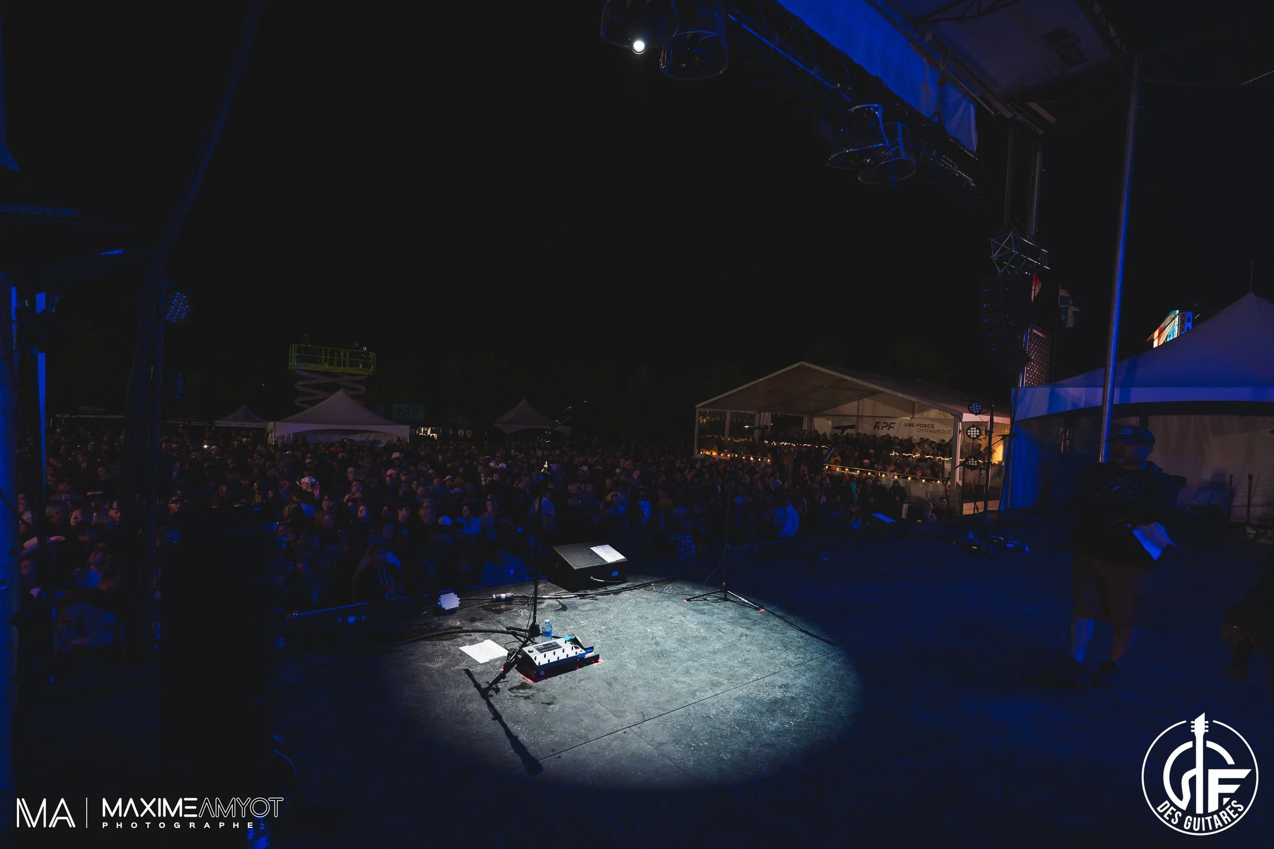 Scène de concert en plein air la nuit avec une foule nombreuse, une scène vide avec des microphones, et un technicien à droite.