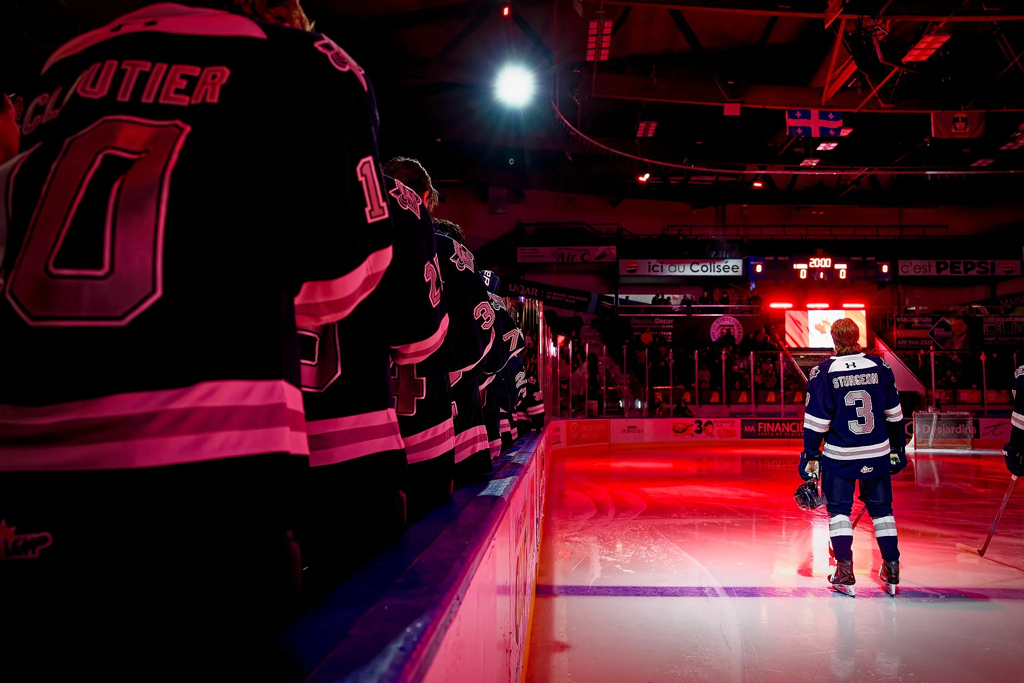 Ice hockey player standing on the ice rink while teammates stand along the bench, with a teammate facing the audience, in an arena with red lighting