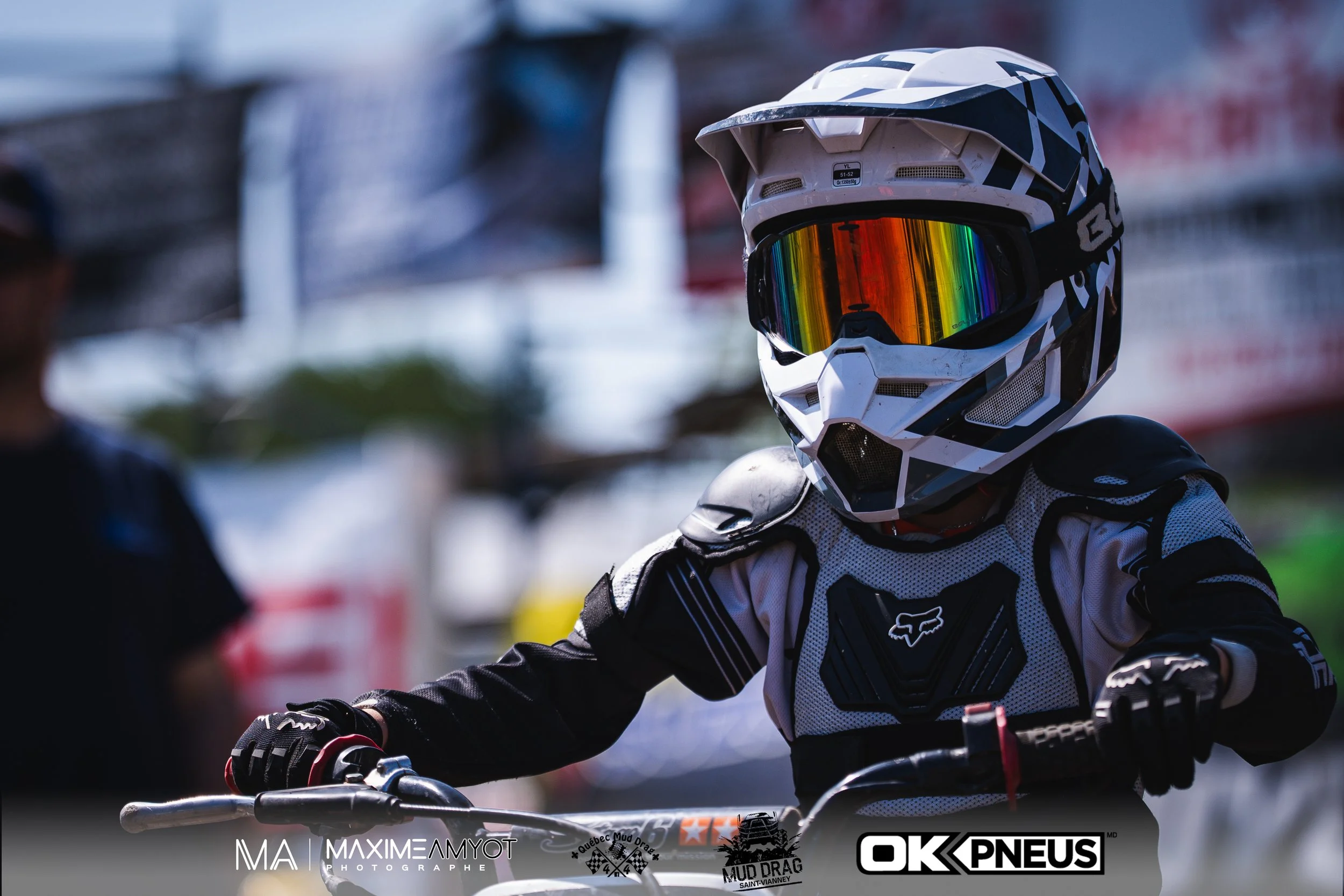 Pilote de motocross en tenue de protection, portant un casque intégral avec visière miroir colorée, sur sa moto lors d'une compétition.
