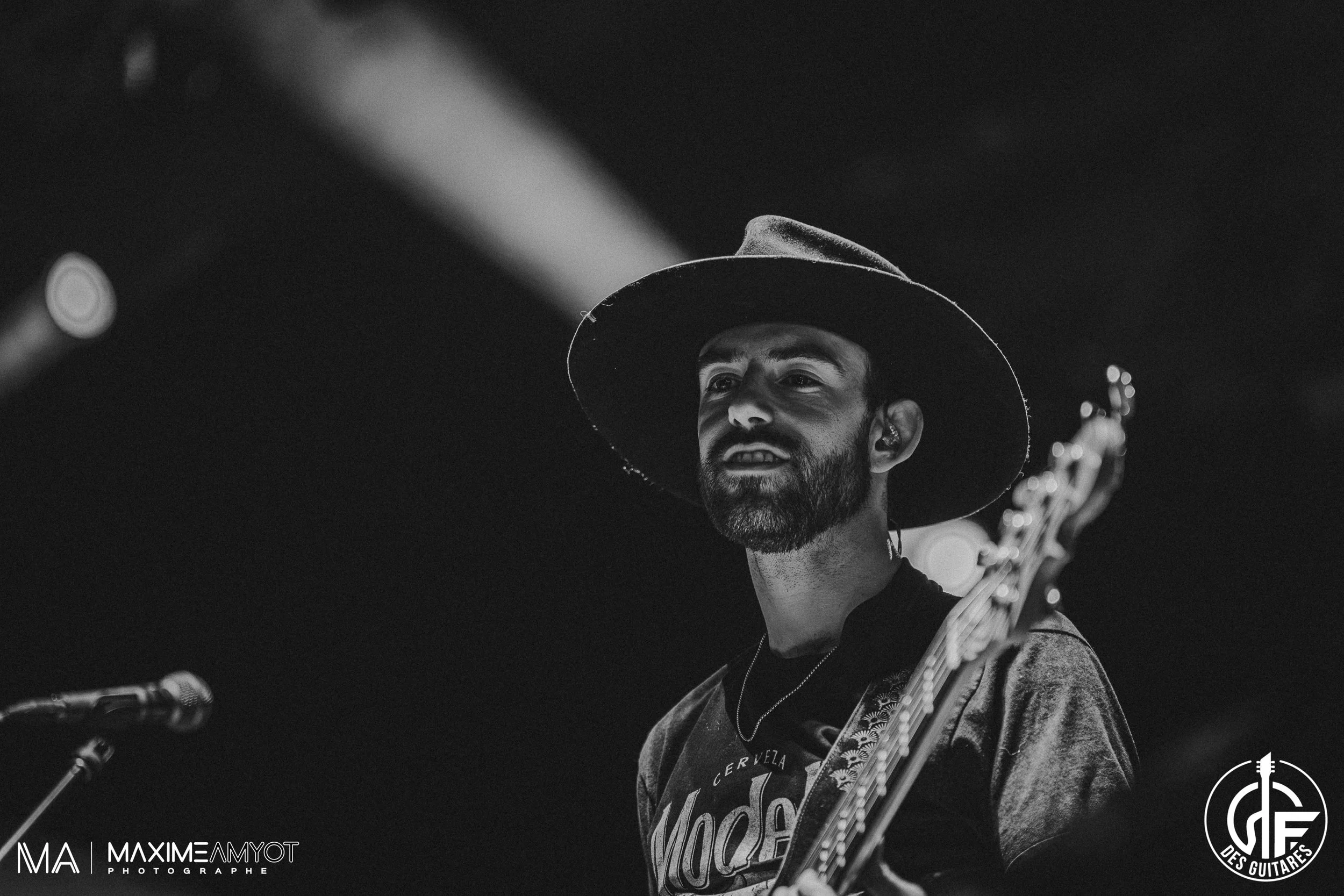 Un homme portant un grand chapeau feutre joue de la guitare lors d'une performance. La photo est en noir et blanc, avec un éclairage provenant de projecteurs au-dessus.